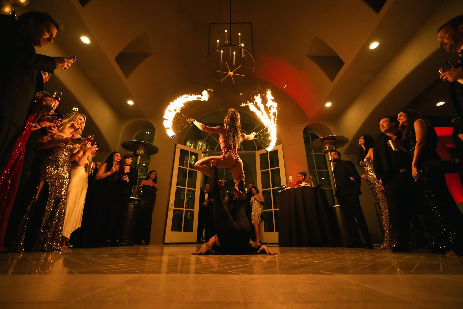 Fire performer spinning flaming props in arc above head during indoor event, surrounded by spectators in formal attire