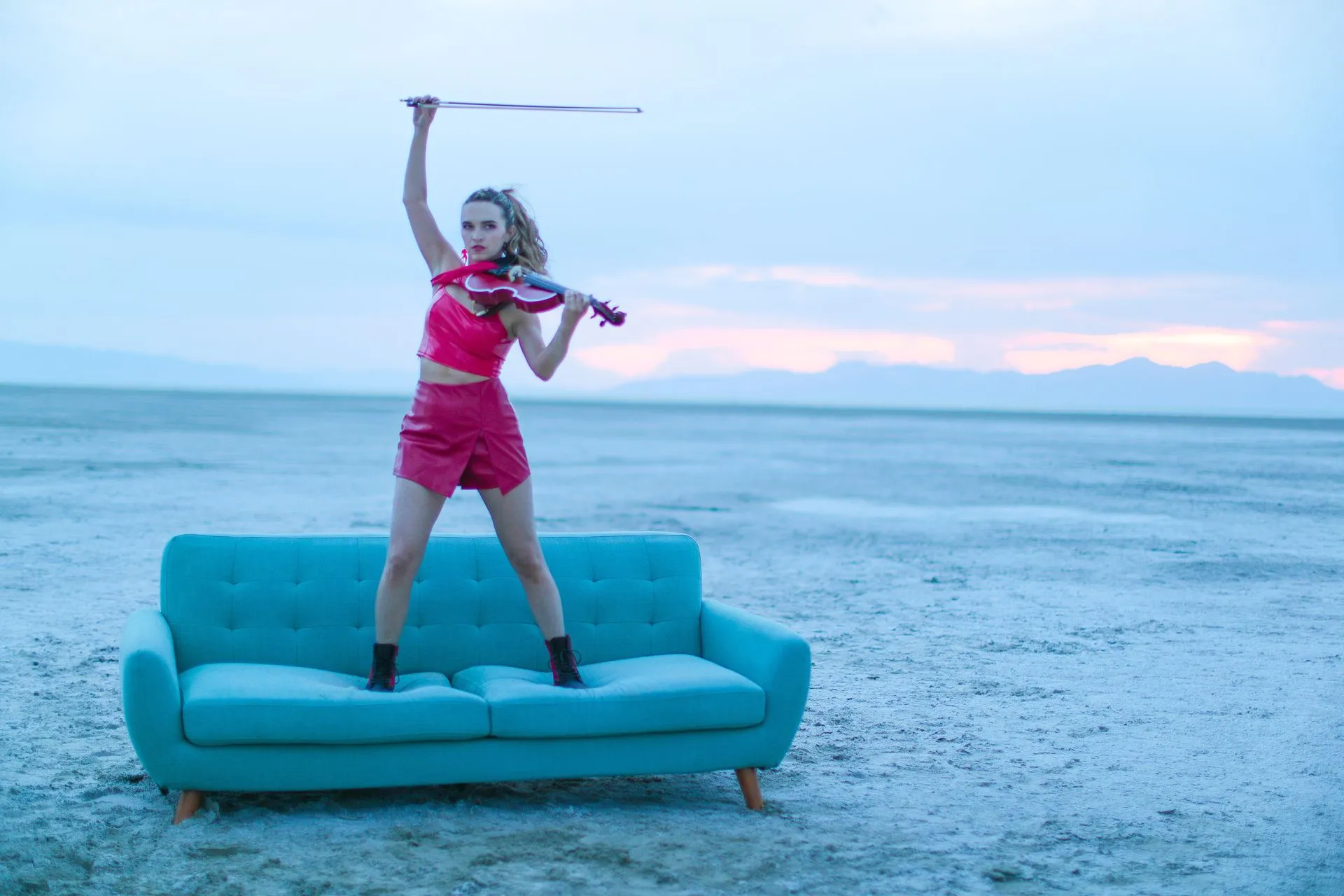 A woman is standing on a blue couch playing a violin.