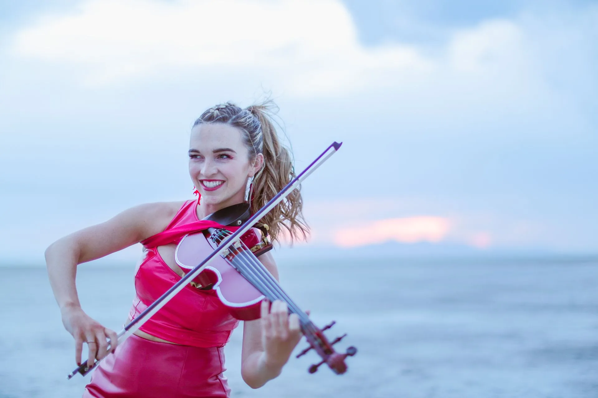 A woman in a red dress is playing a violin on the beach.