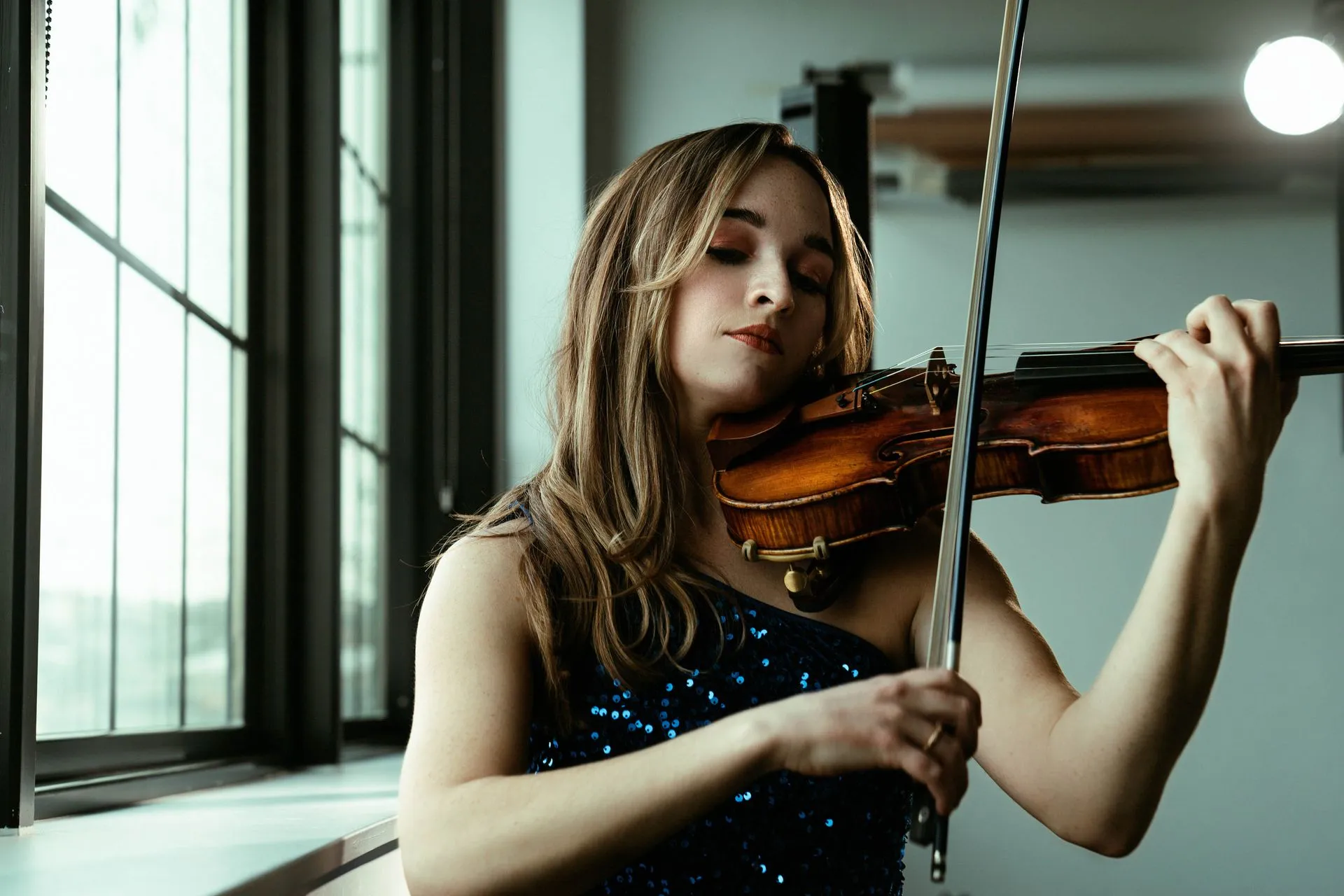A woman in a blue dress is playing a violin in front of a window.