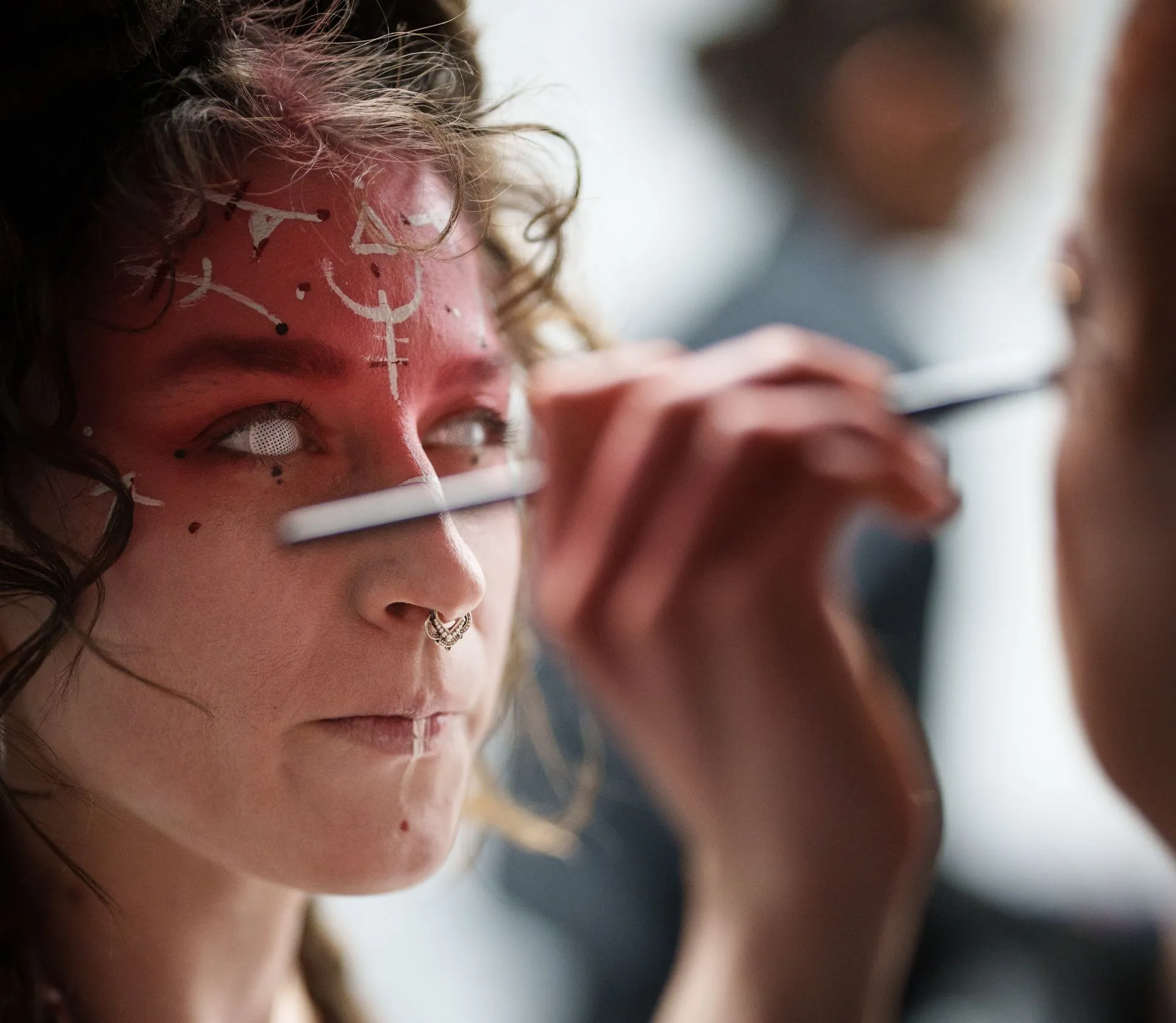 Person with white eyes and red face paint being worked on with a makeup brush, nose ring visible.