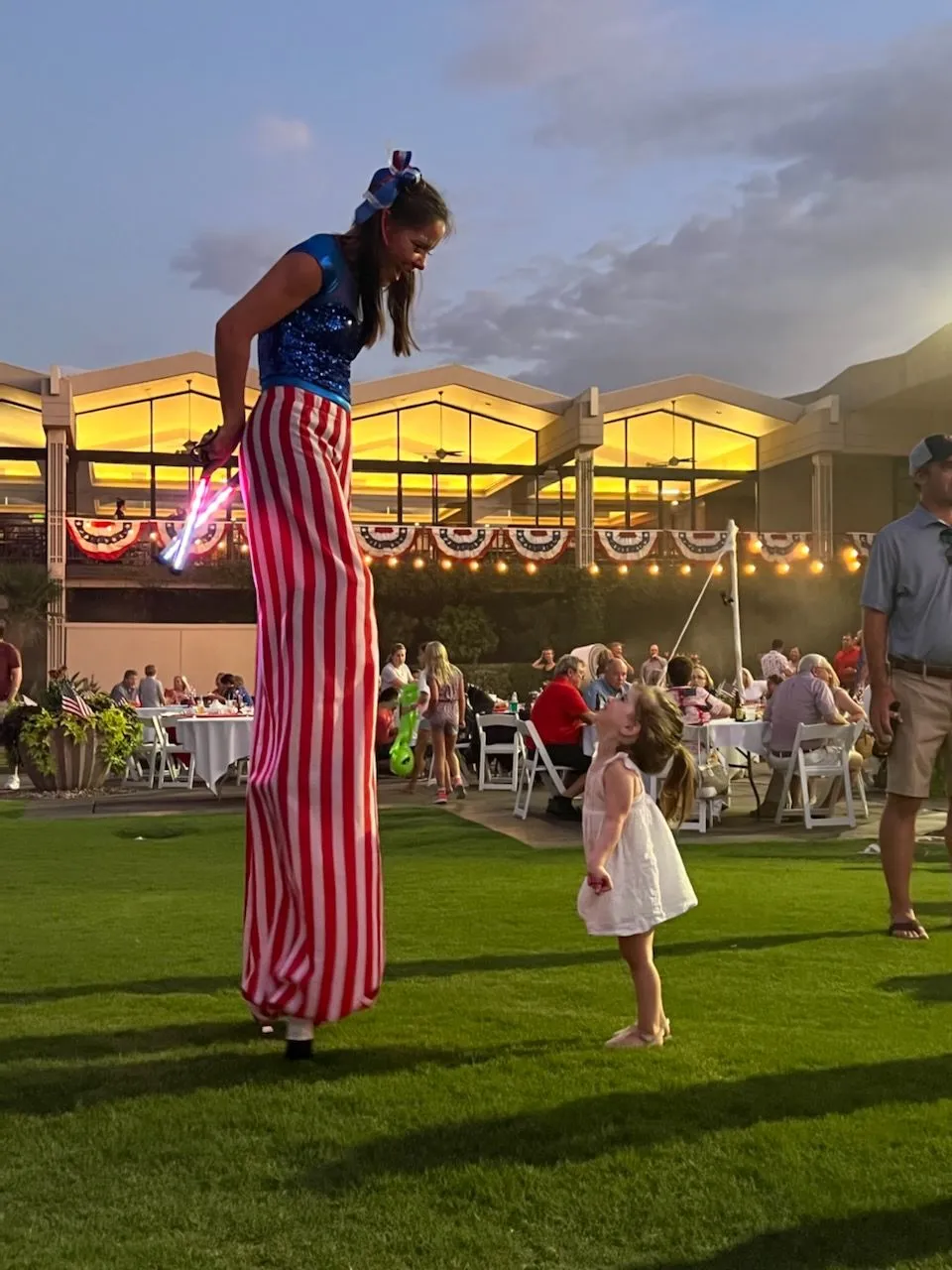 A little girl is standing next to a woman on stilts.
