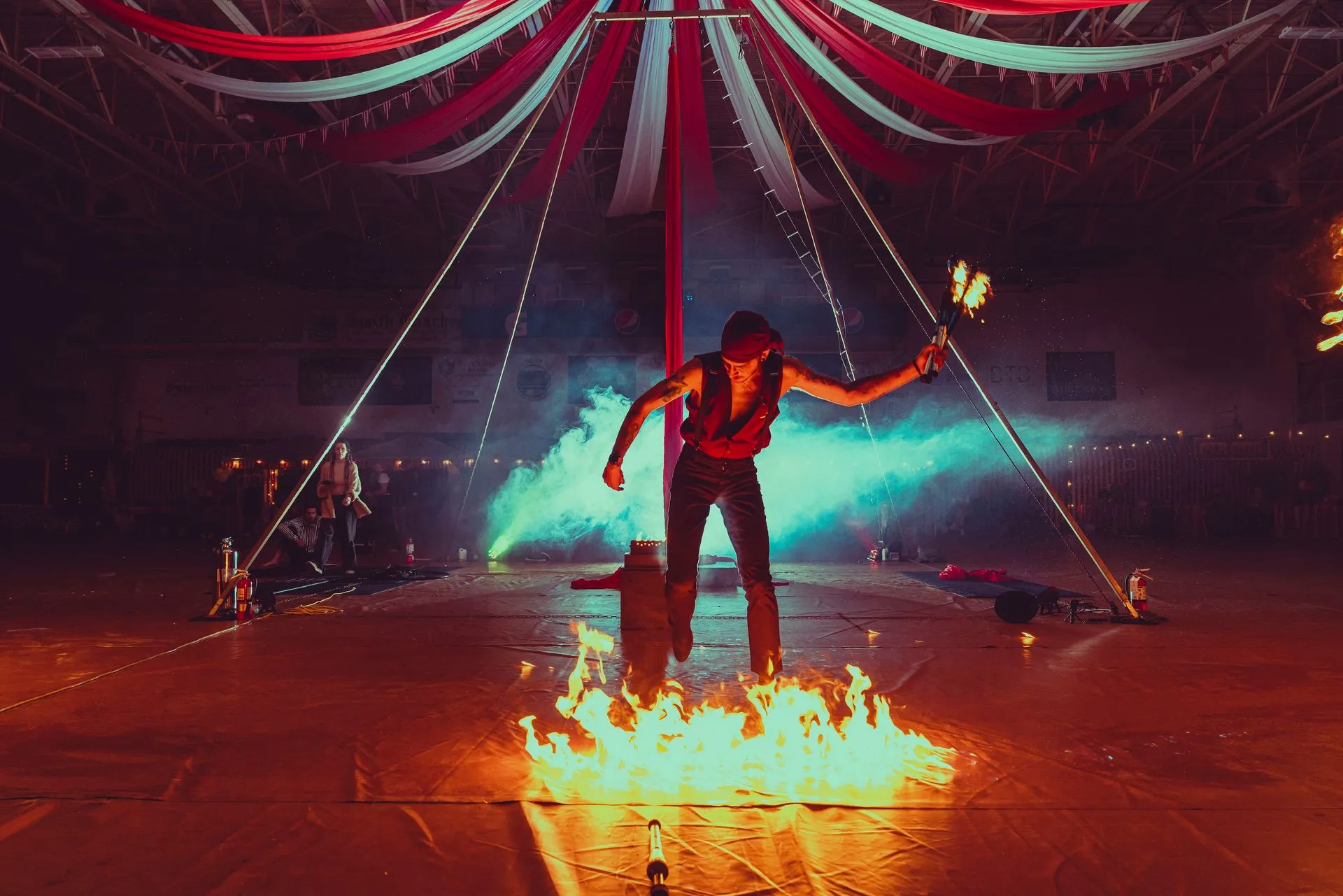A man is performing aerial acrobatics in a circus tent with fire coming out of his hands.