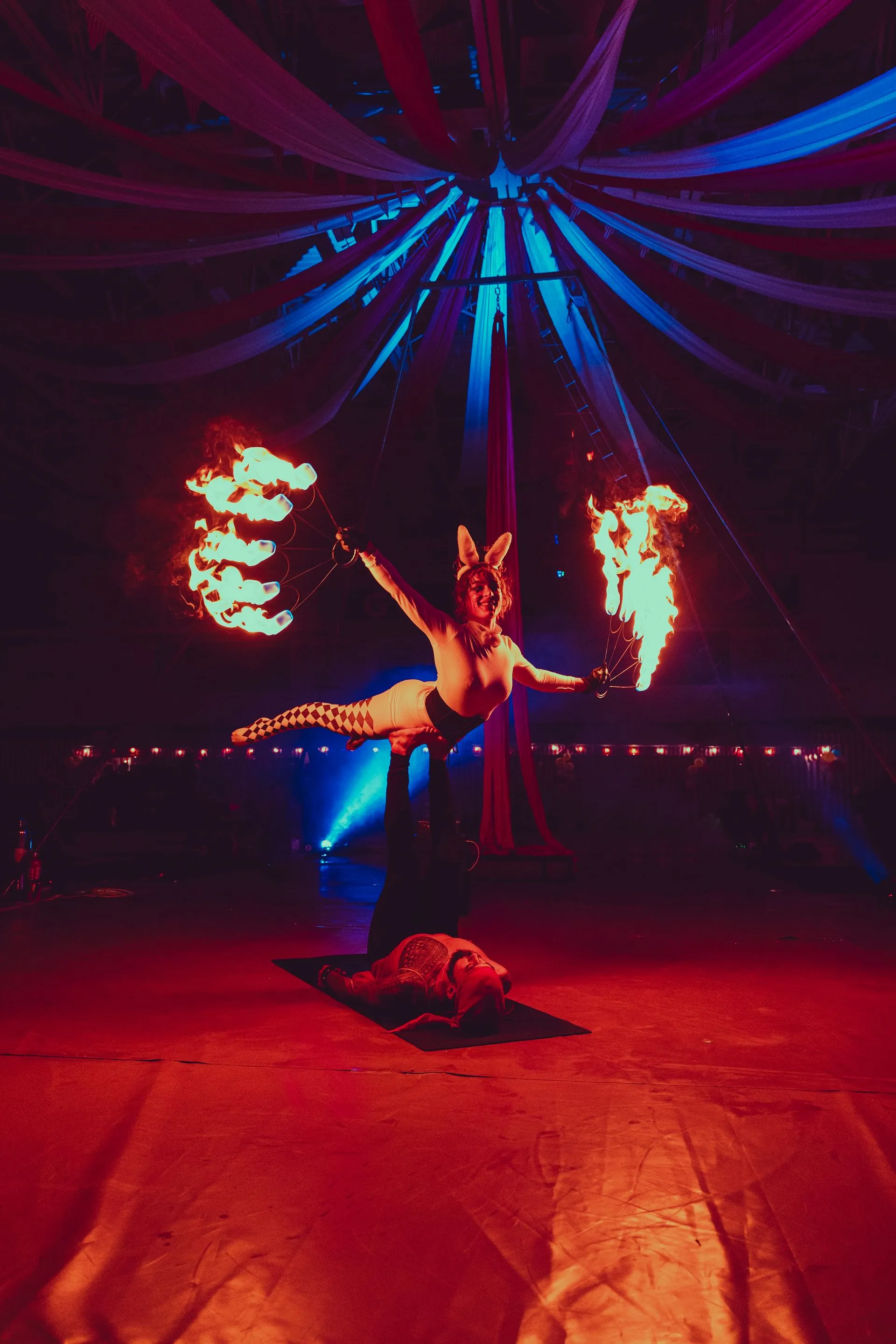 A woman is performing aerial acrobatics in a circus tent with fire coming out of her hands.