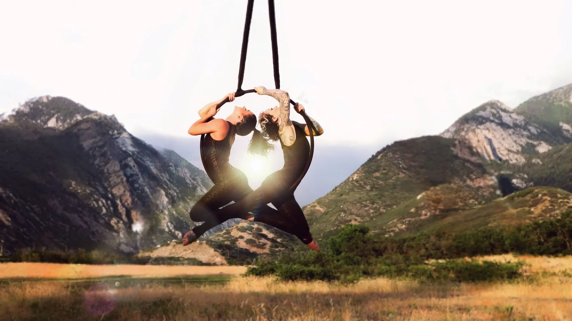 Two people are hanging from a rope in a field with mountains in the background.