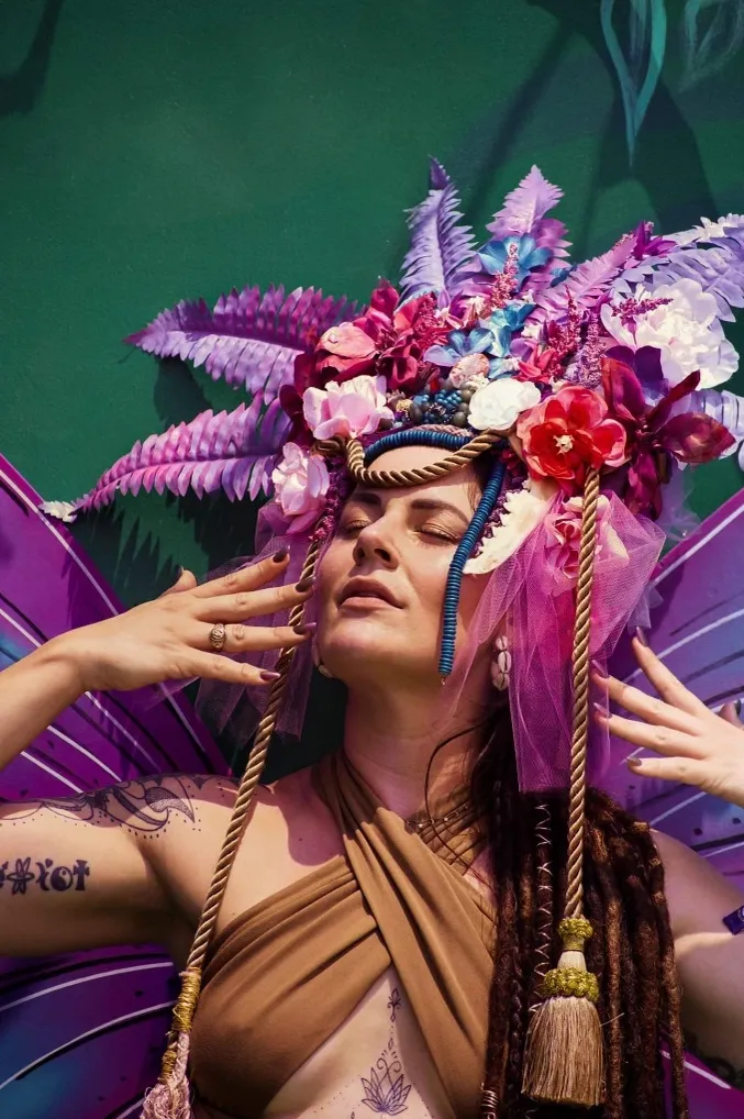 A woman wearing a crown of flowers and wings is standing in front of a green wall.