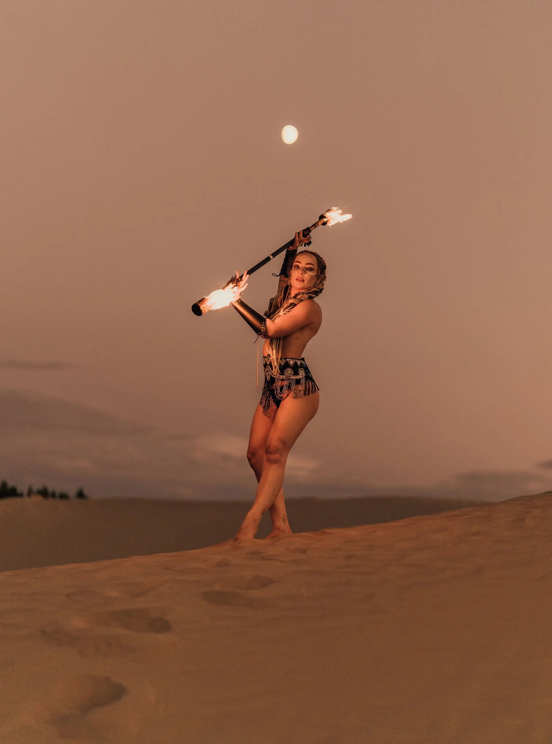 A woman is standing on top of a sand dune holding a fire stick.