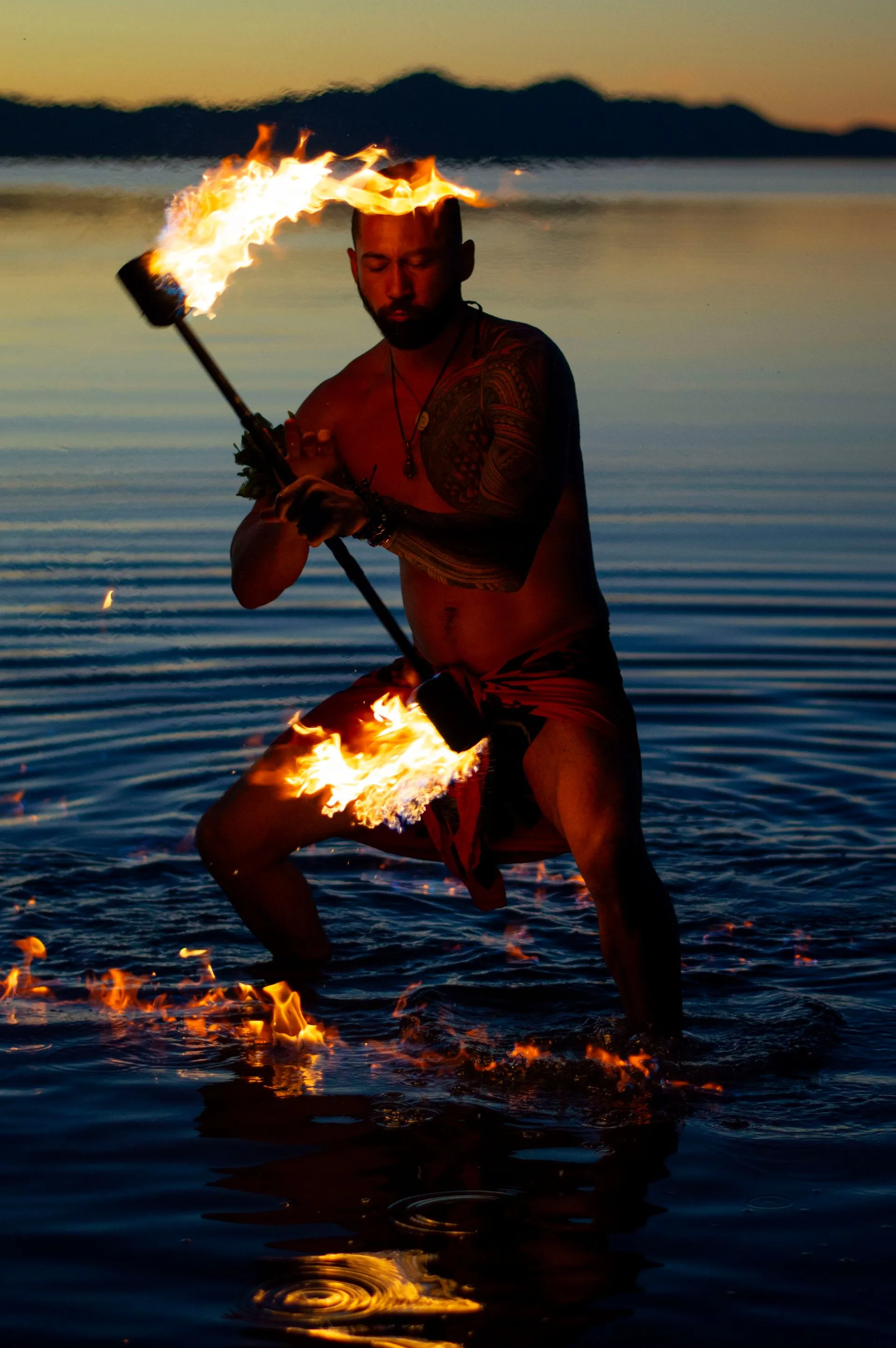 Fire dancer in water, holding flaming staff. Fire around head and legs, sunset in background.
