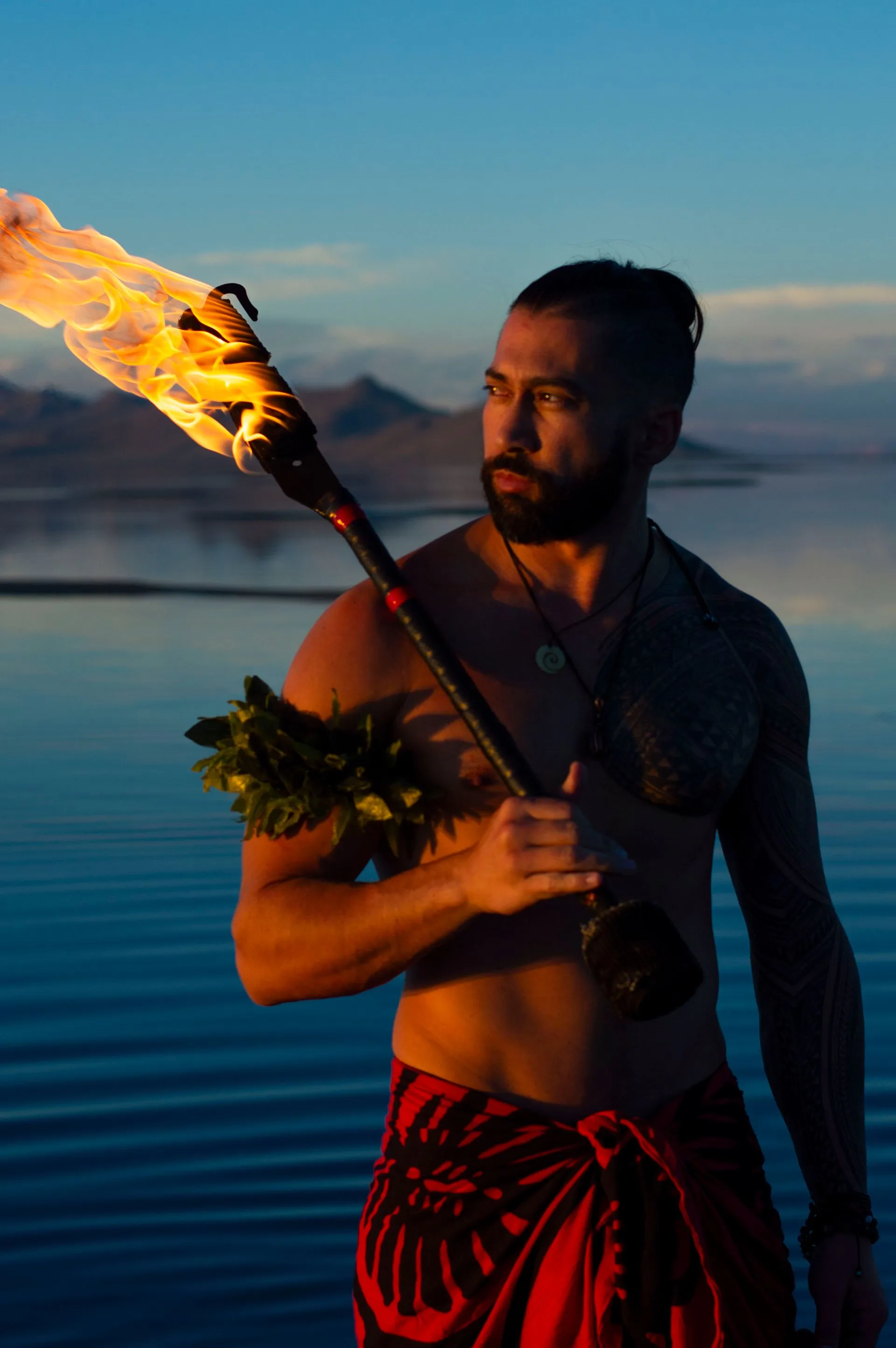 Man holding a flaming torch, wrapped in a red sarong. Standing in water with mountains in the background.
