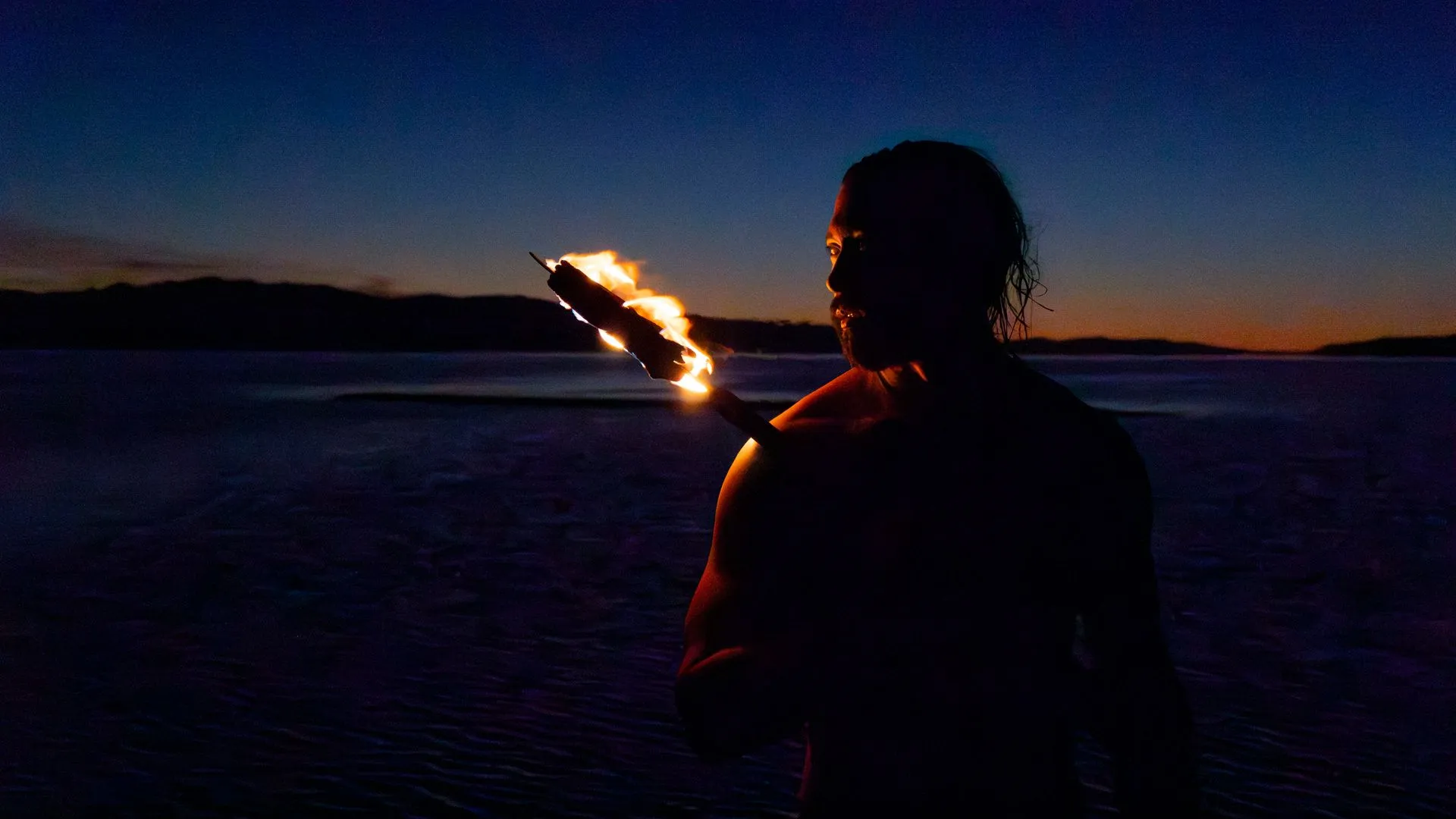 Person holding a torch, silhouetted against a dark, waterside landscape at dusk.