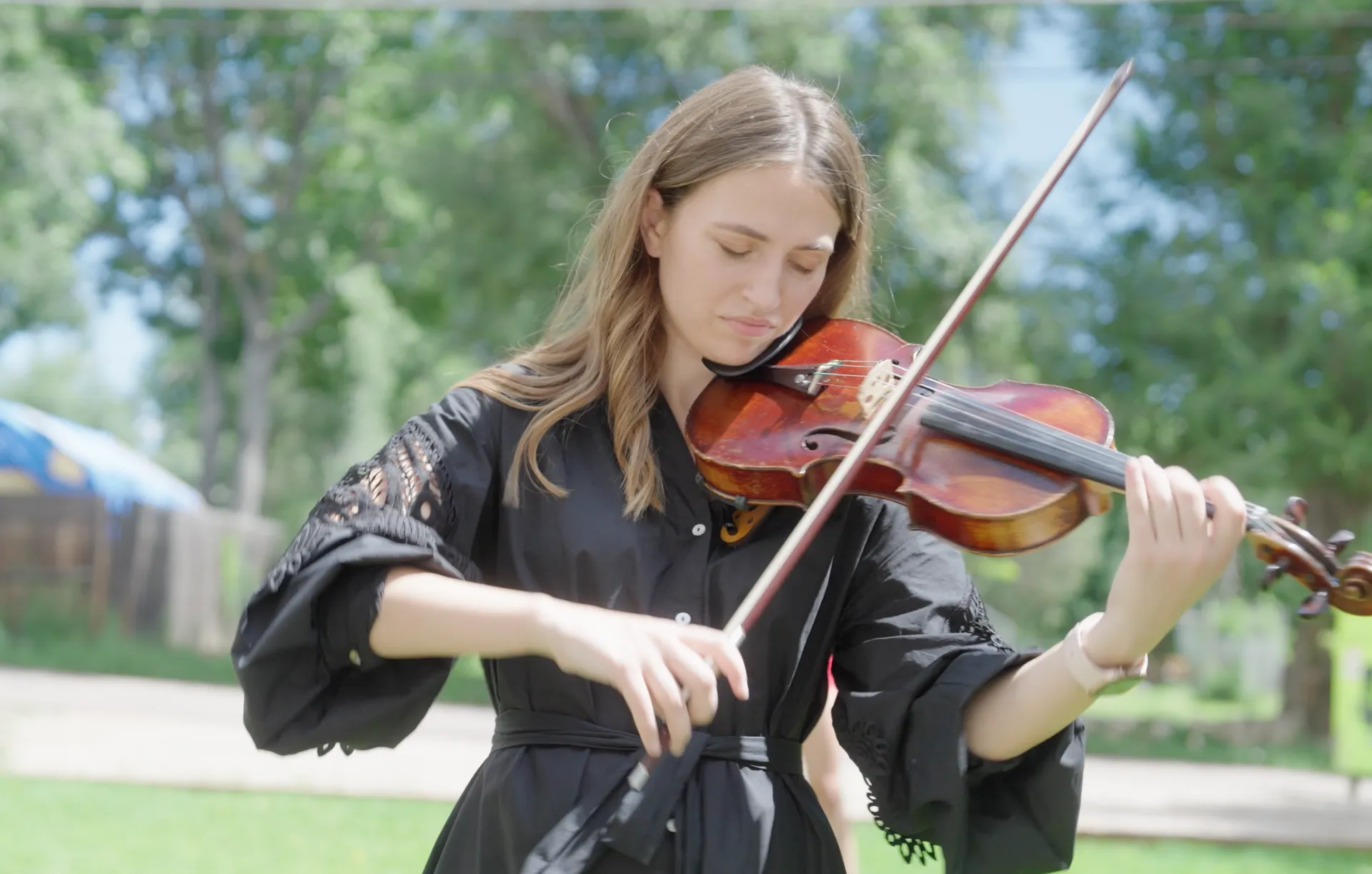 A woman in a black dress is playing a violin in a park.