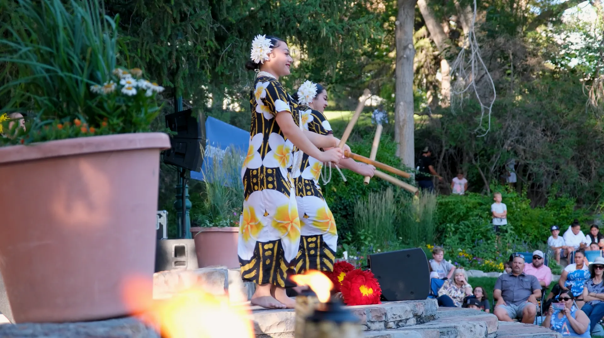 Two women are dancing hula in front of a crowd in a park.