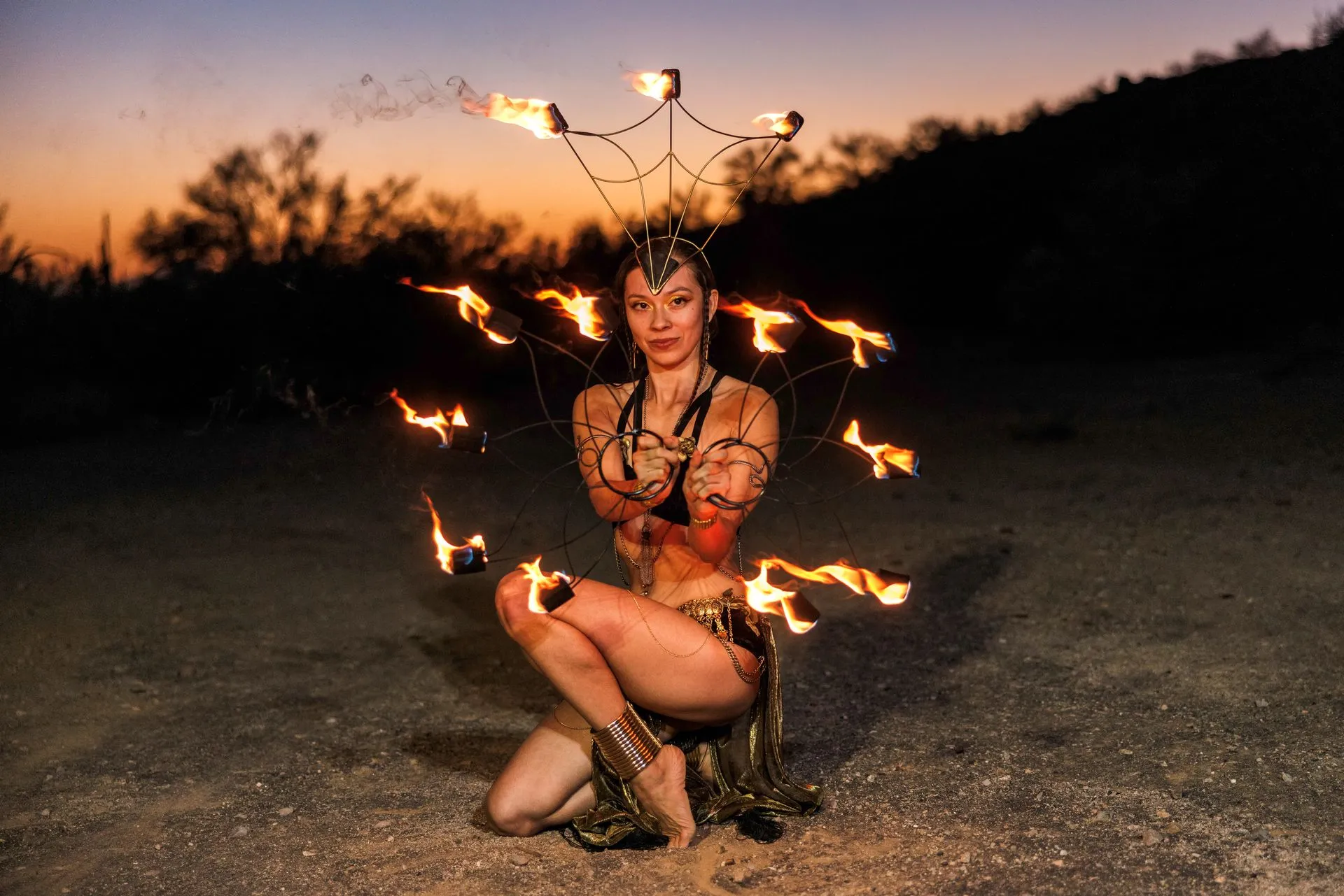 A woman is kneeling down in the dirt holding a fan of fire.