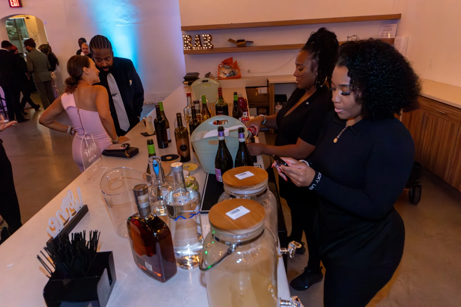 Two people in black stand behind a long bar counter lined with liquor bottles & drink dispensers under a lit BAR marquee sign while guests stand nearby