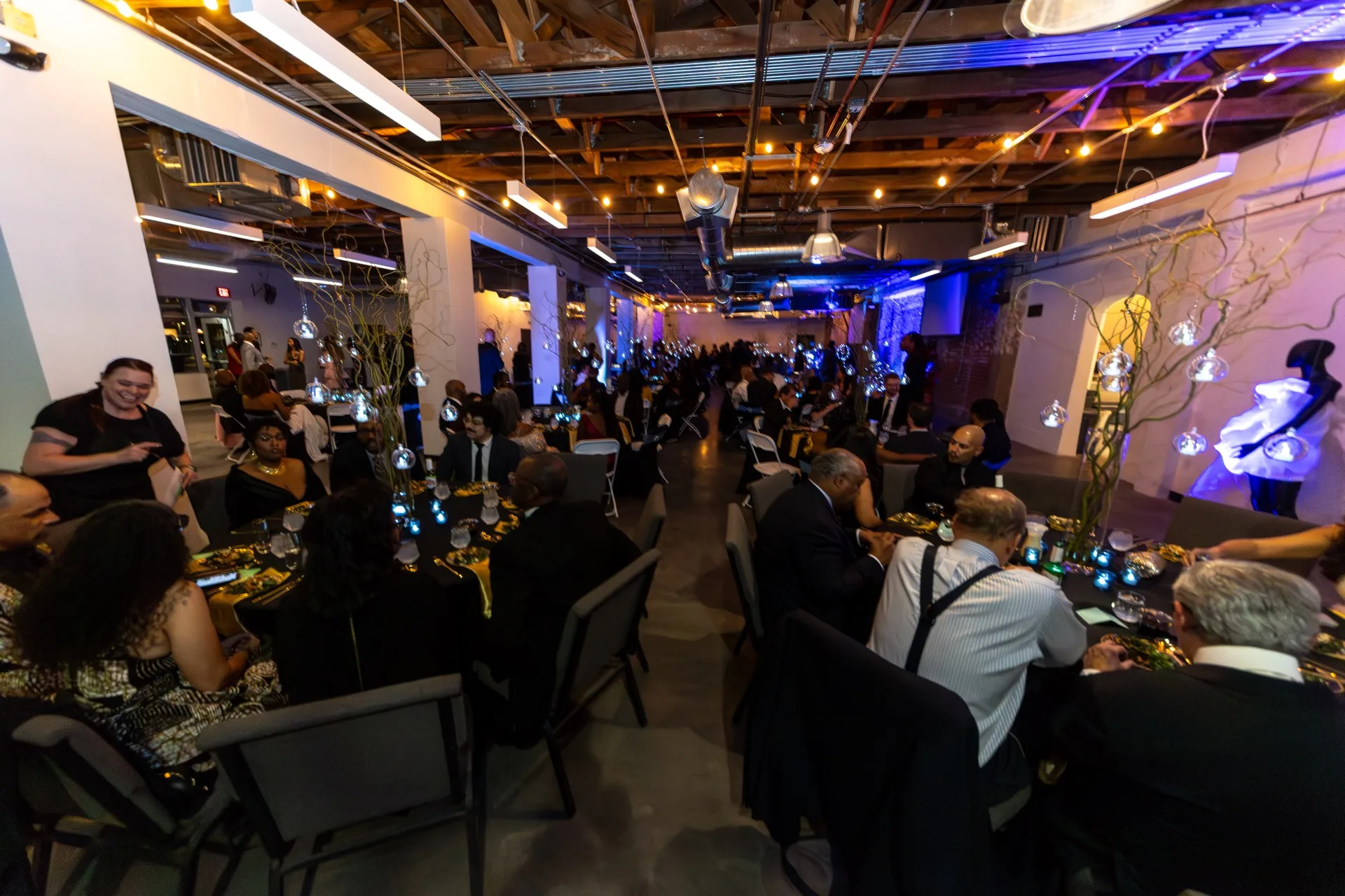 Guests dine at long tables under warm hanging ceiling lights, blue wall uplighting, and bulb-branch centerpieces in an event hall.