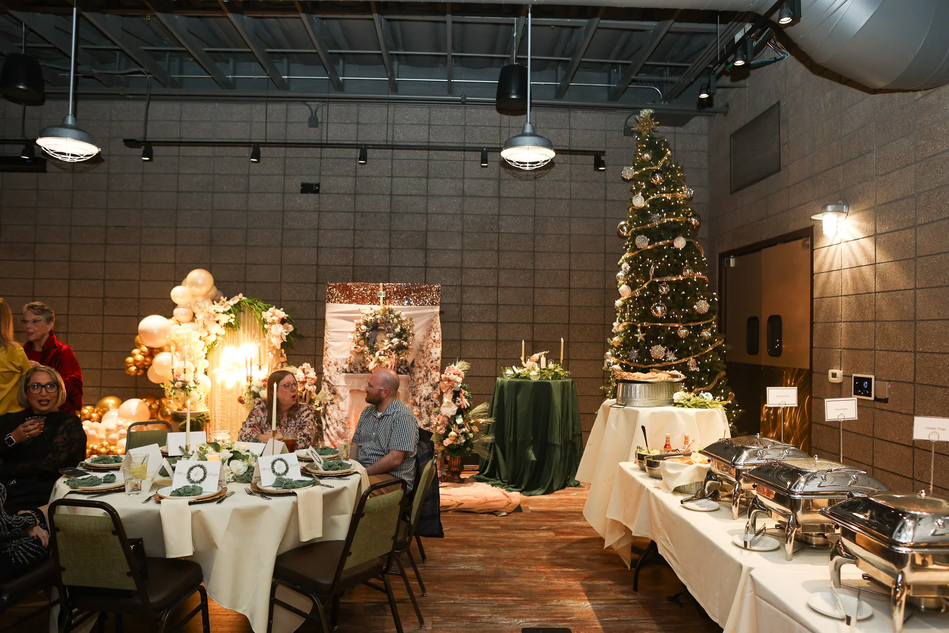 Banquet room with balloon and floral arch, decorated tree, round tables set with linens and candles, chafing dishes on a side table.