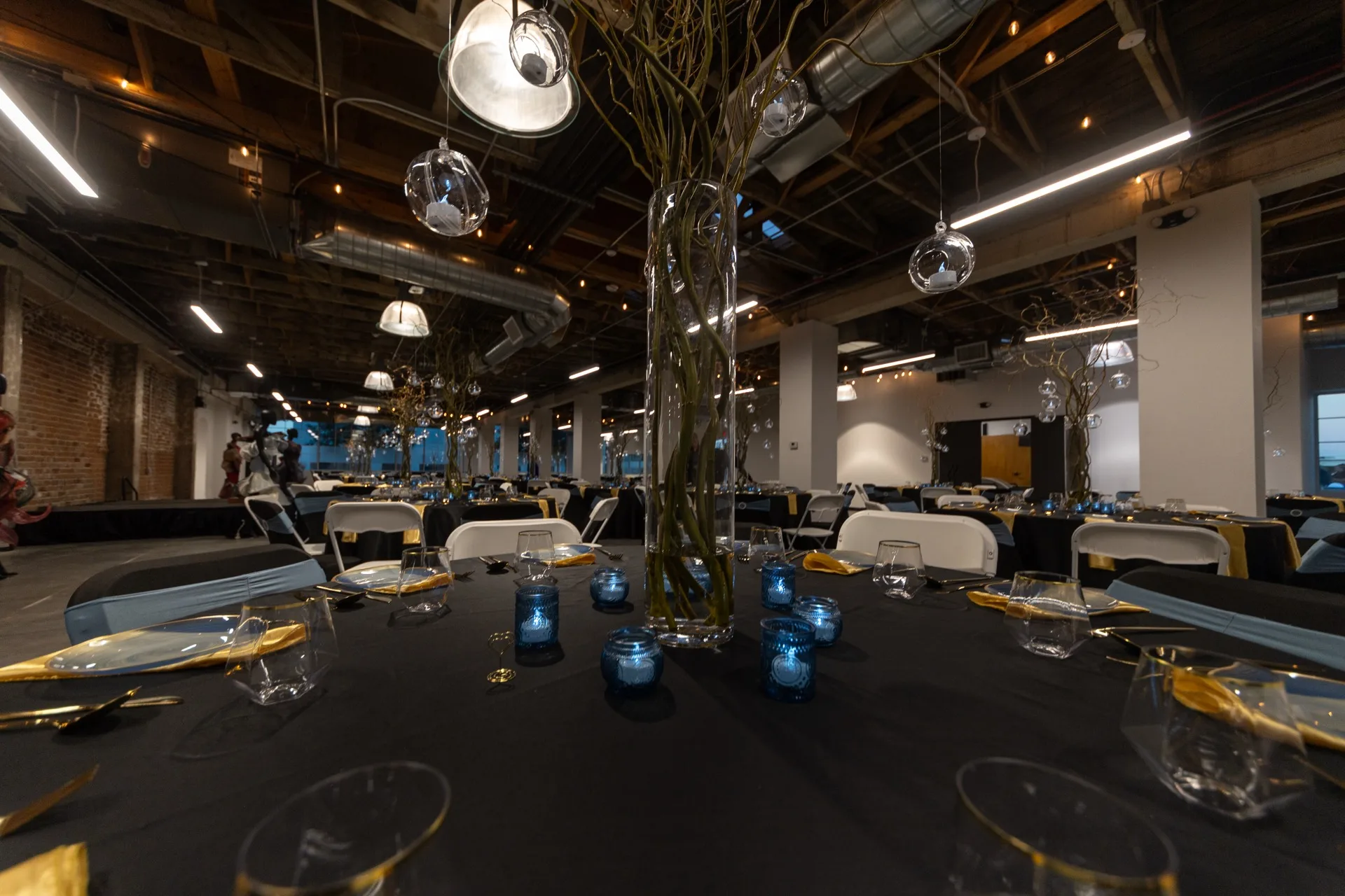 Modern event space with black tablecloths, gold accents, blue candlelit centerpieces, and geometric hanging lights in an industrial setting.