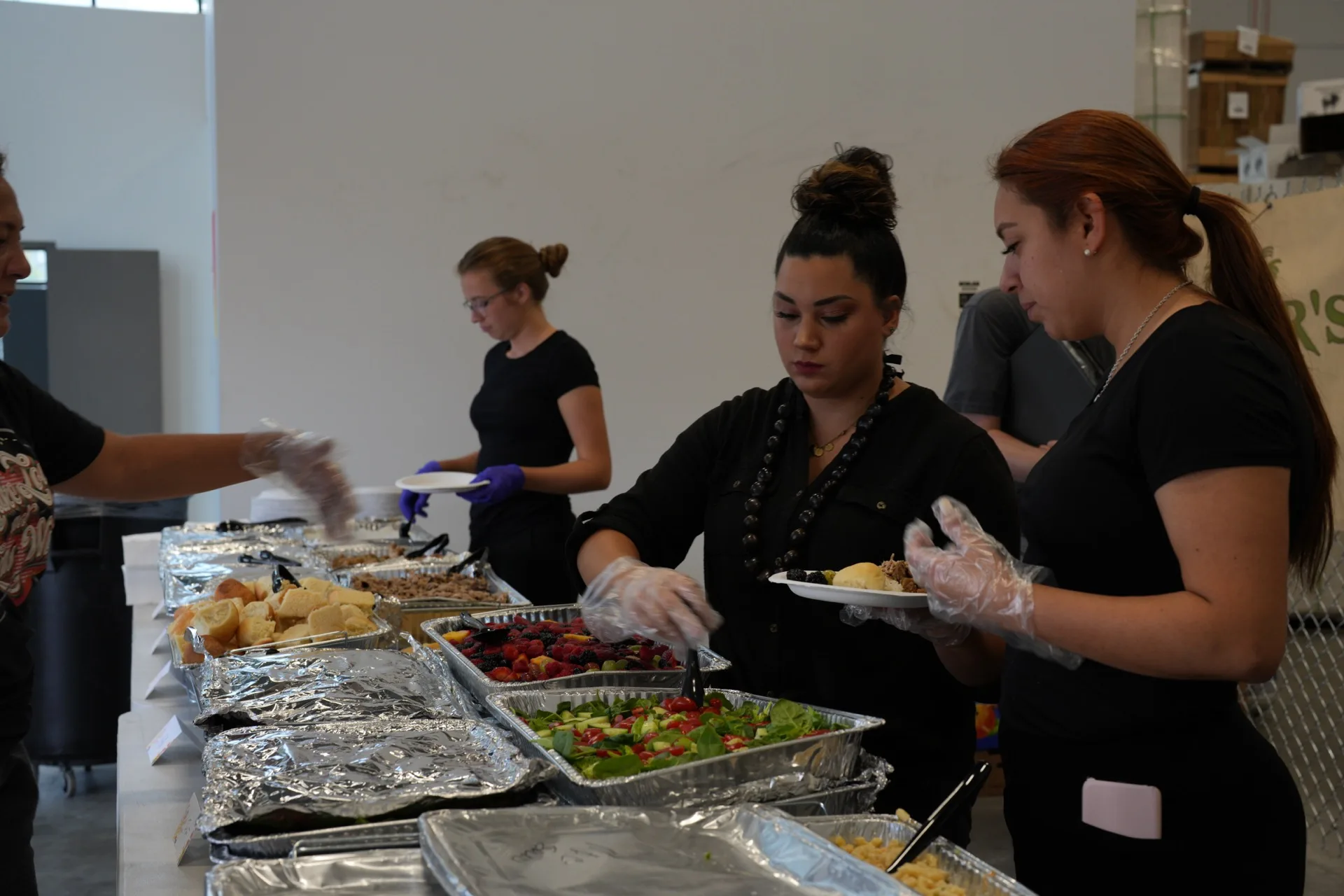Staff in black uniforms and white gloves serve catered foods including salads, berries, and chips from aluminum trays.