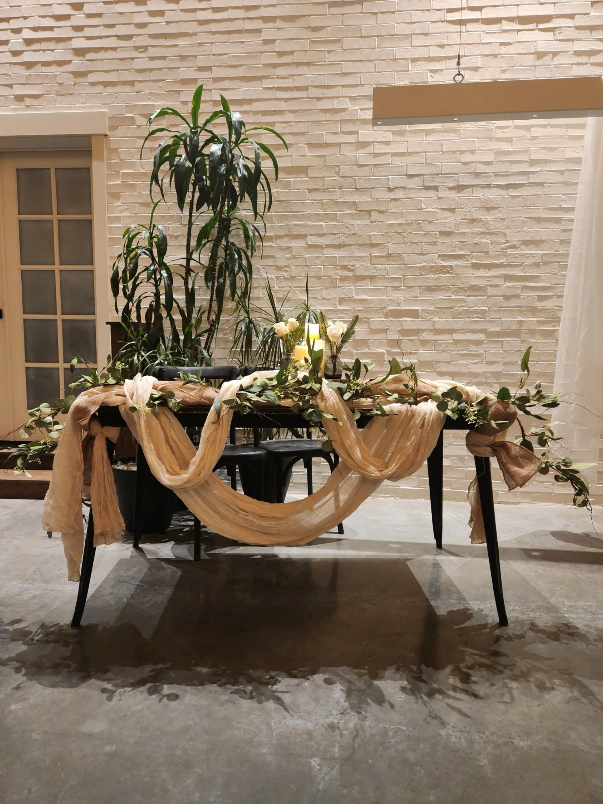 A dark wood table draped with tan fabric swags and greenery garland, candles and white flowers on top, against a brick wall.