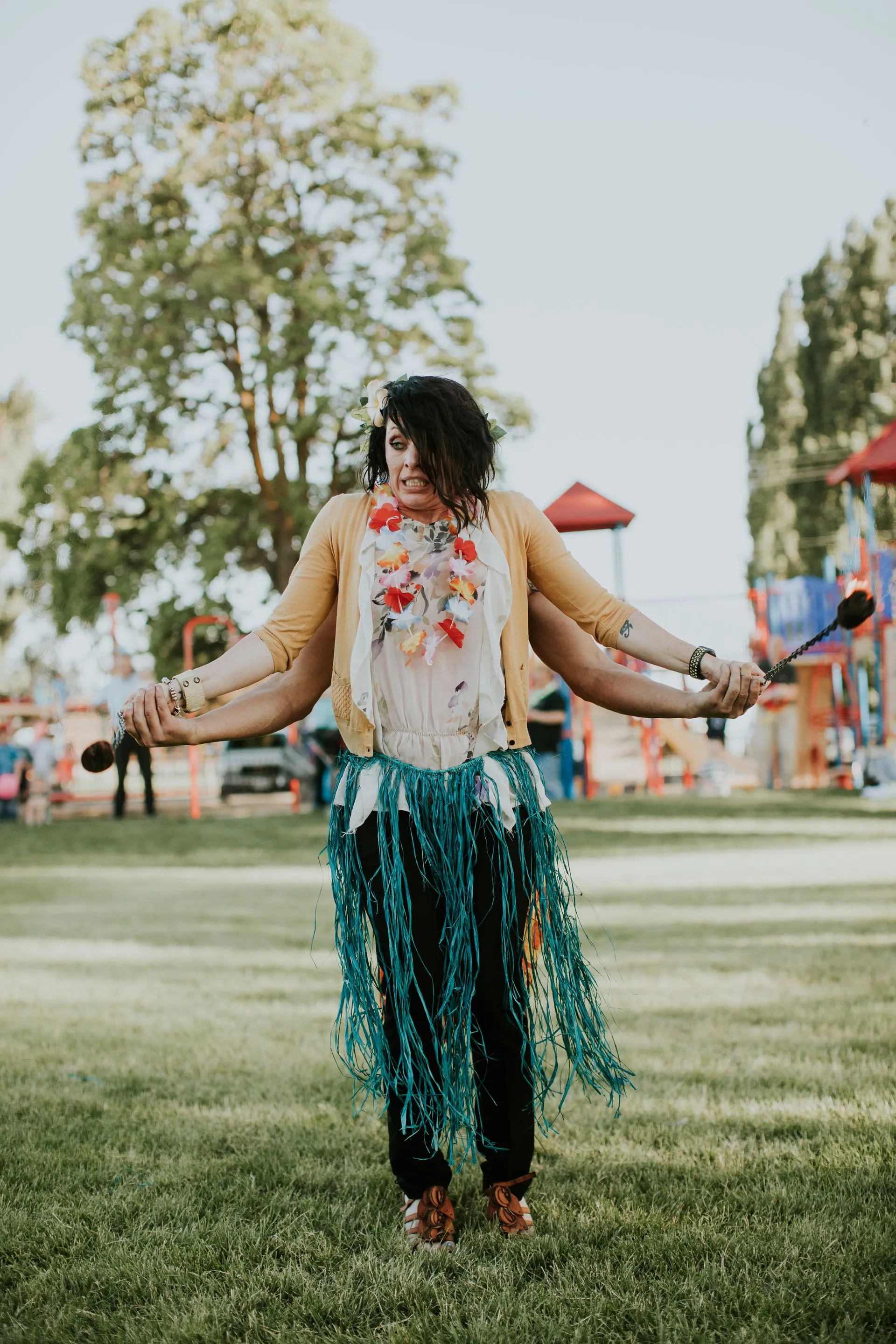 A woman in a hula skirt is standing in the grass with her arms outstretched.
