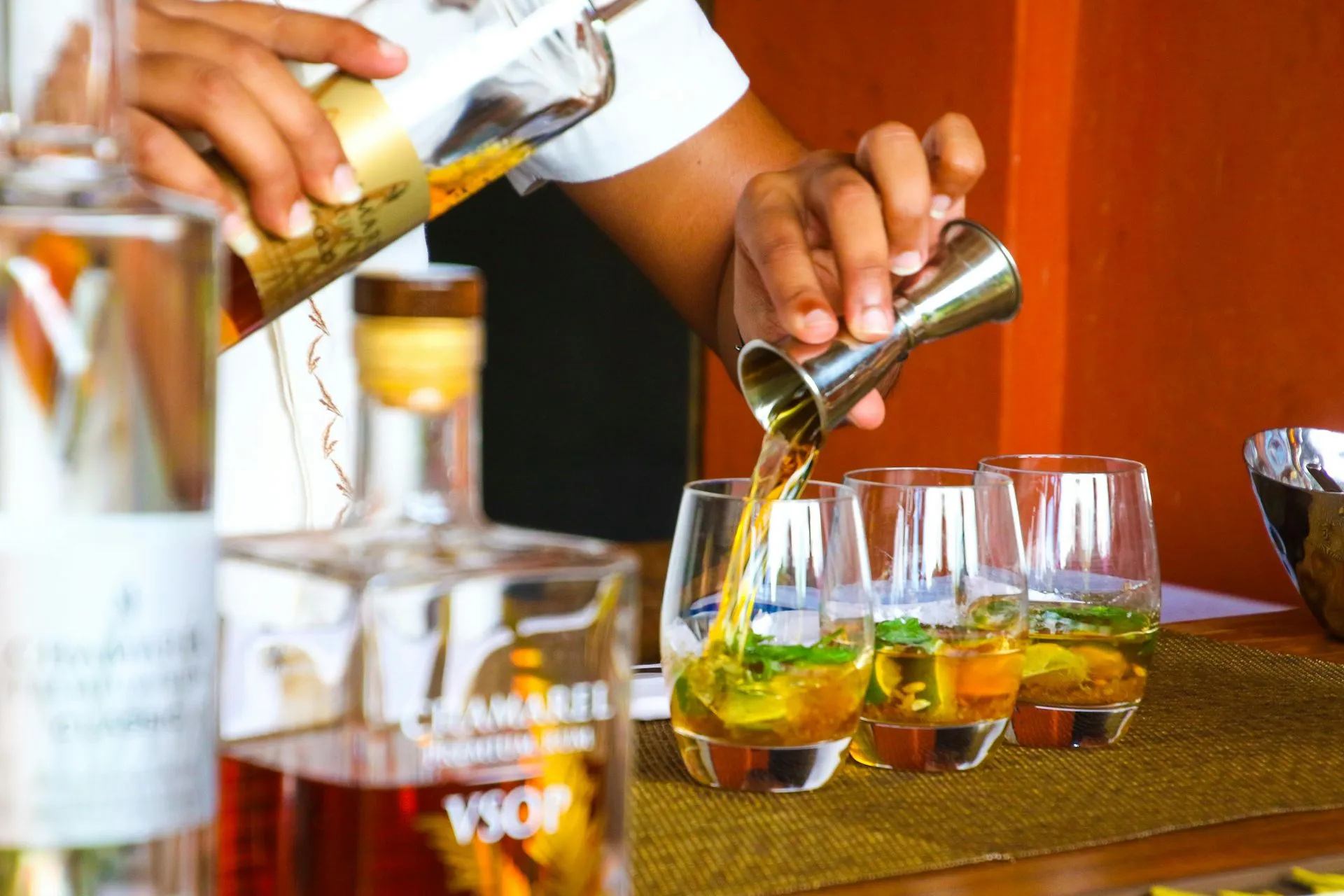 Bartender pouring amber liquor from a jigger into three glasses with mint garnish on a bar.
