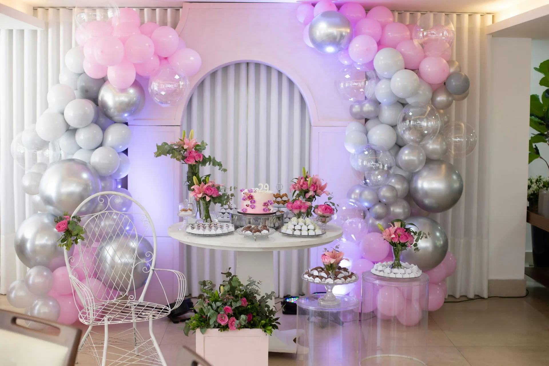 Party decorations: pink, silver, and white balloons frame a dessert table with flowers and cake.