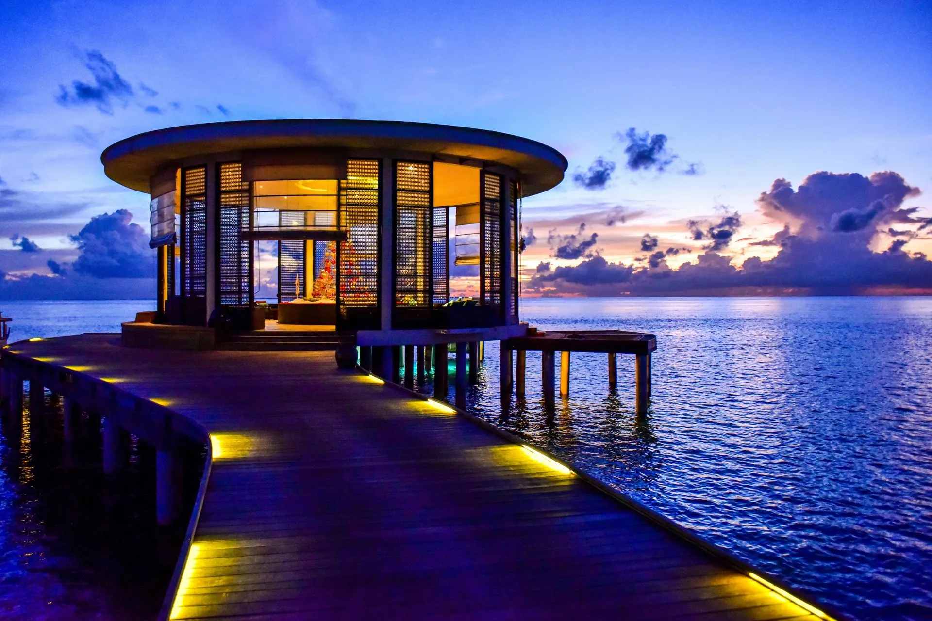 Wooden pier leads to a circular building over ocean at sunset. The building has glass walls, lit interior.