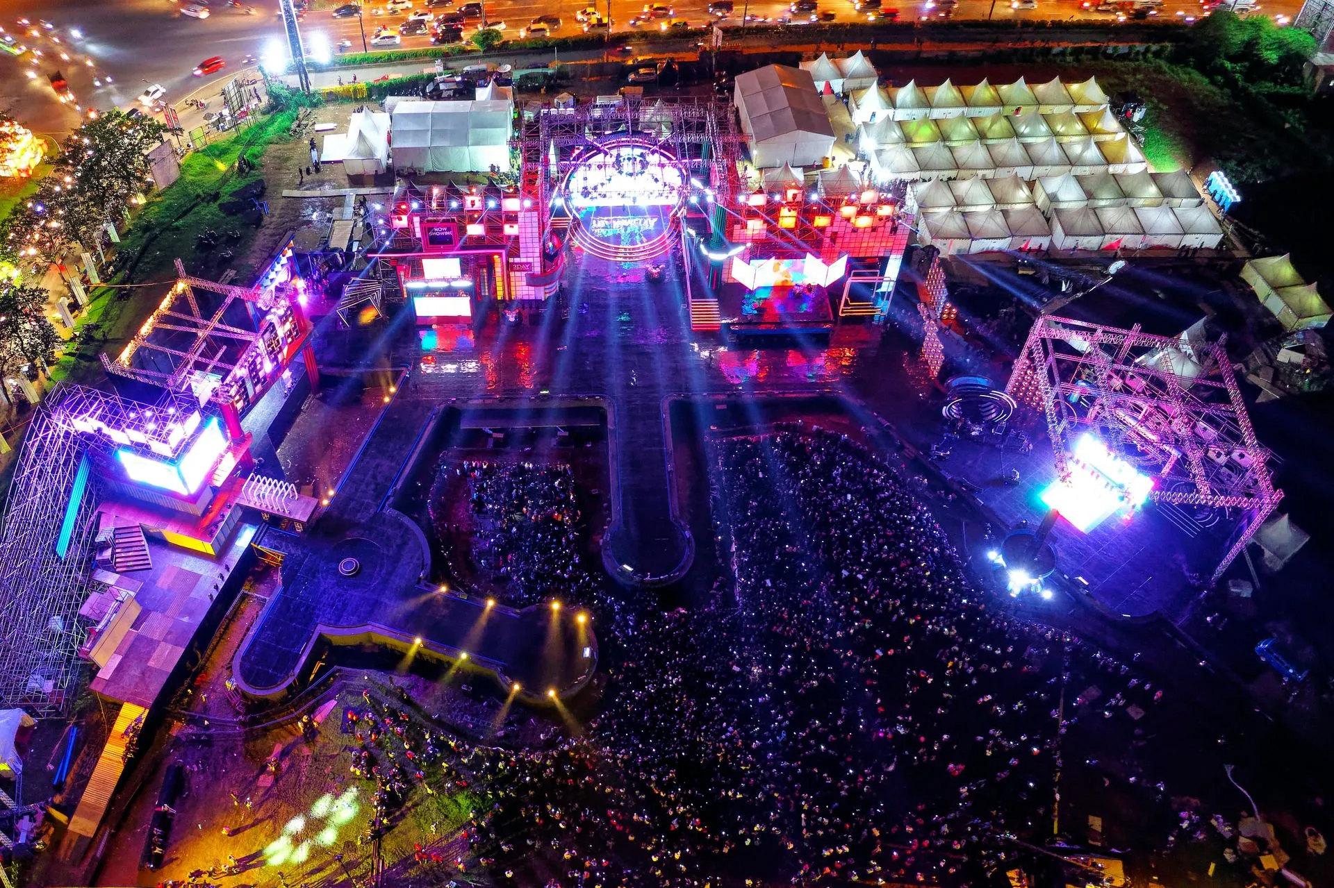 Aerial view of a brightly lit outdoor concert with a large crowd and stages glowing with colorful lights.