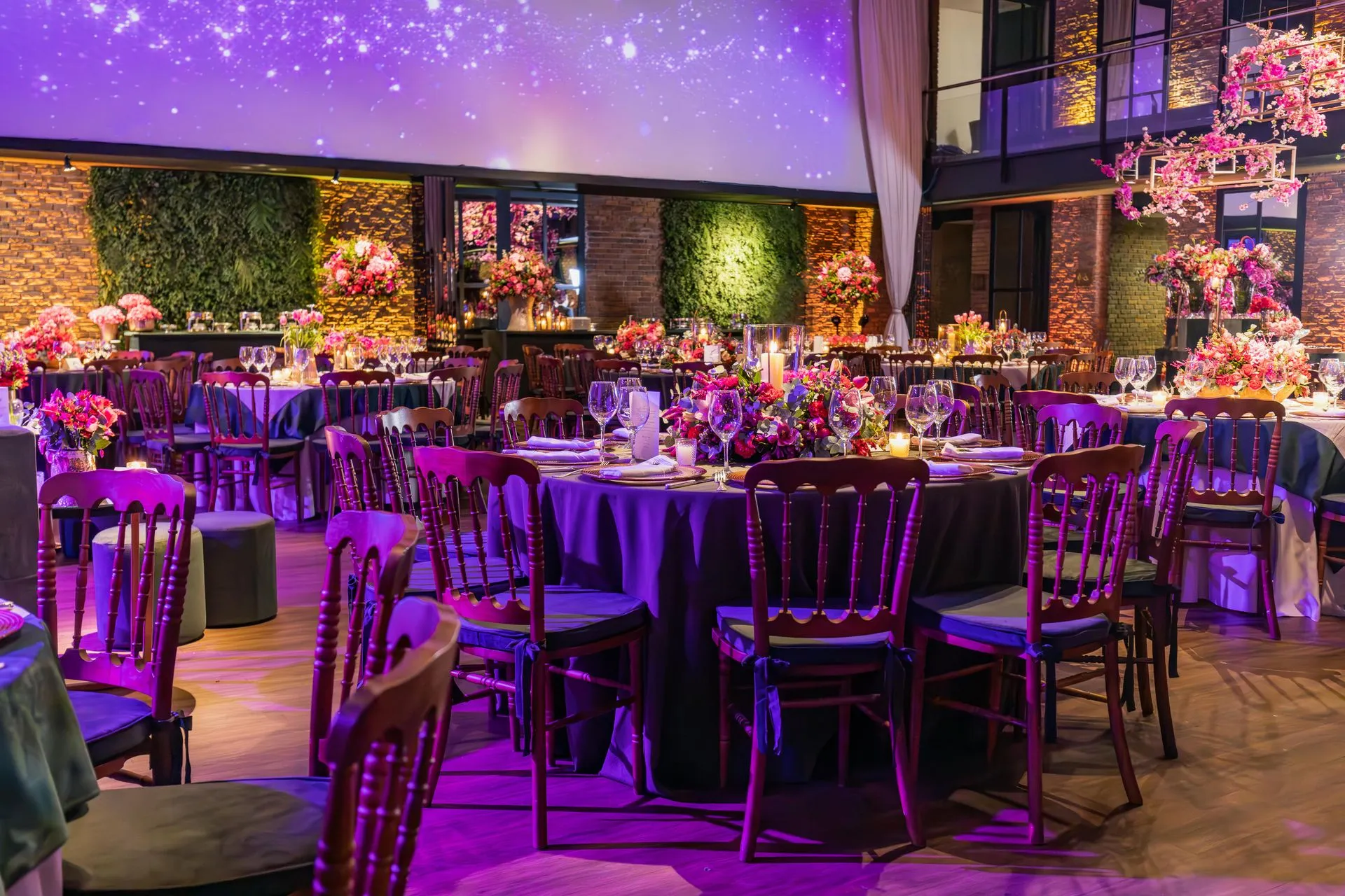 Elegant event hall with tables set for a party; violet and pink lighting, floral centerpieces, wooden chairs, brick walls.