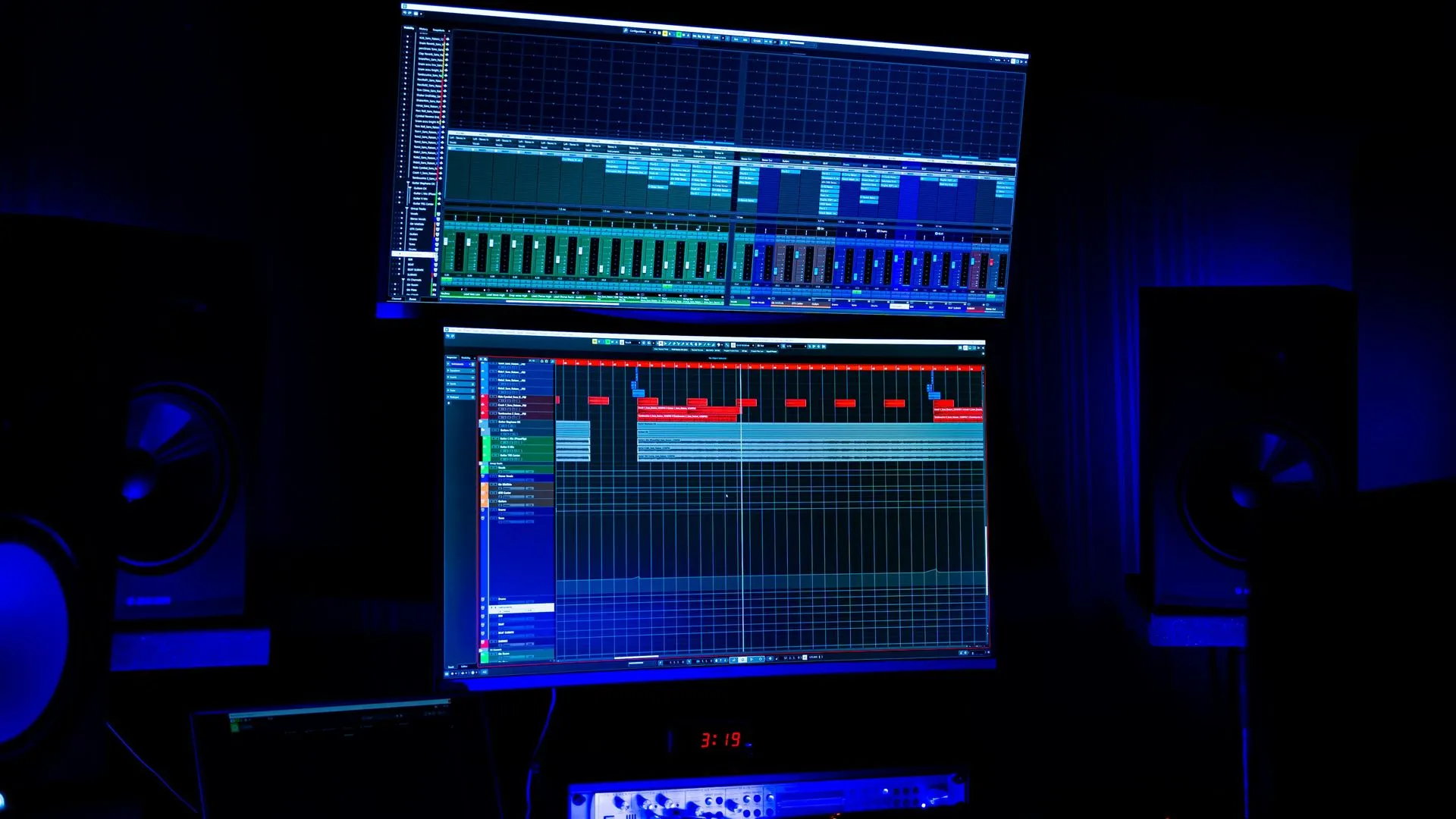 Dark recording studio with two stacked monitors displaying audio editing software. Blue lighting illuminates the space.