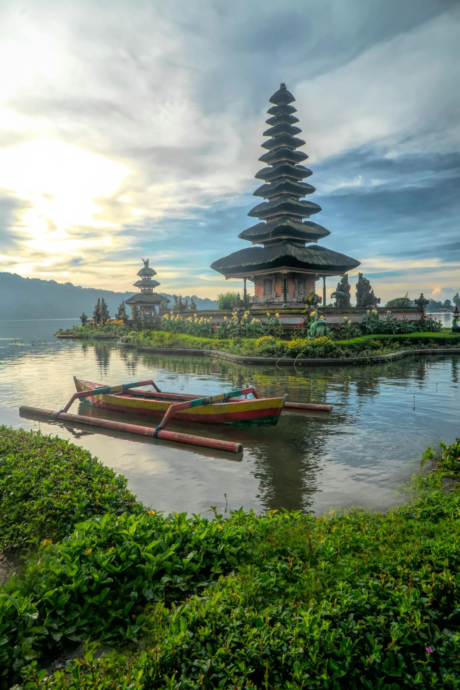 Pura Ulun Danu Beratan temple in Bali, Indonesia, on a lake. A traditional boat sits in front, surrounded by green foliage.
