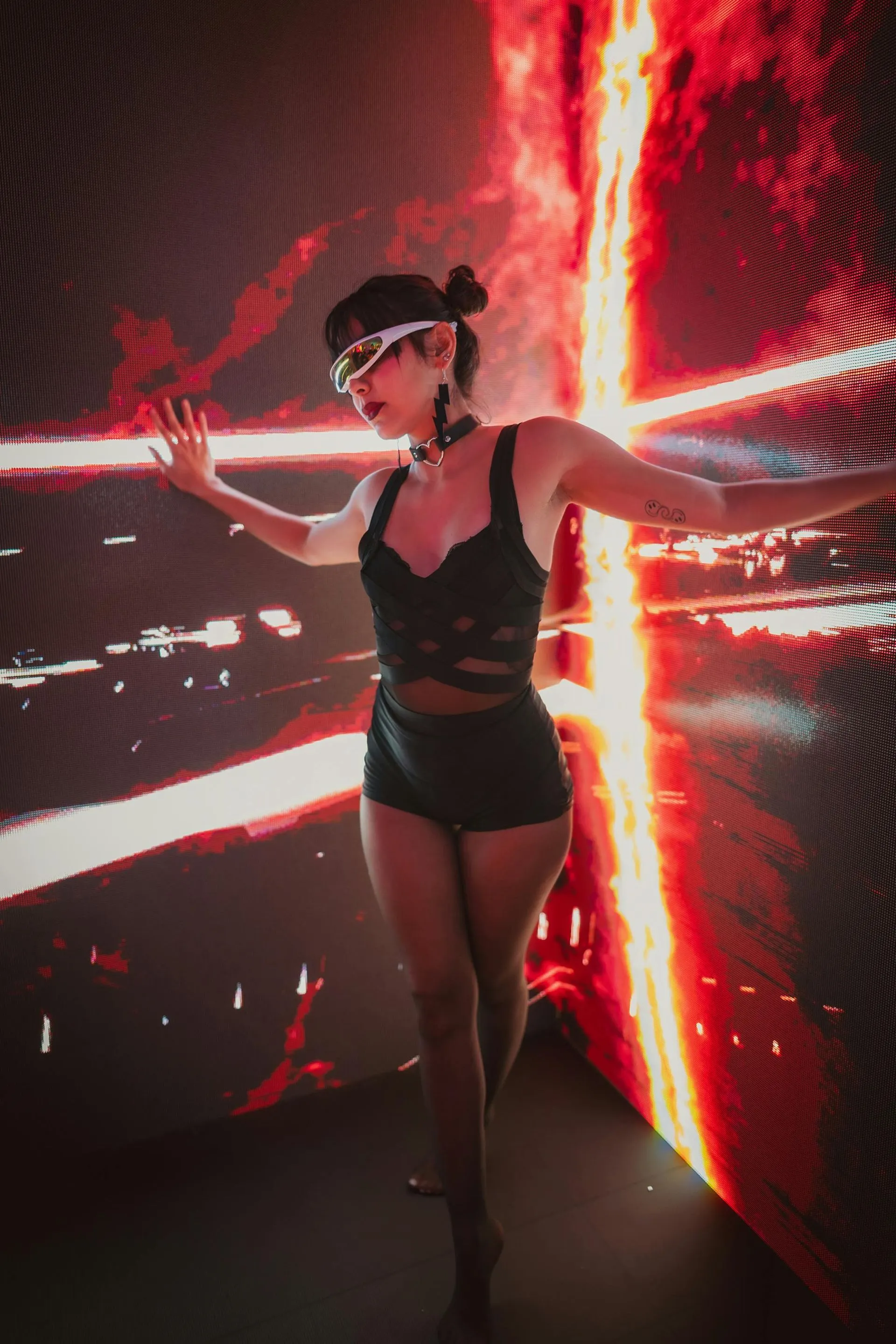 Woman in lingerie, sunglasses, and choker, arms outstretched in a red, glowing corridor.