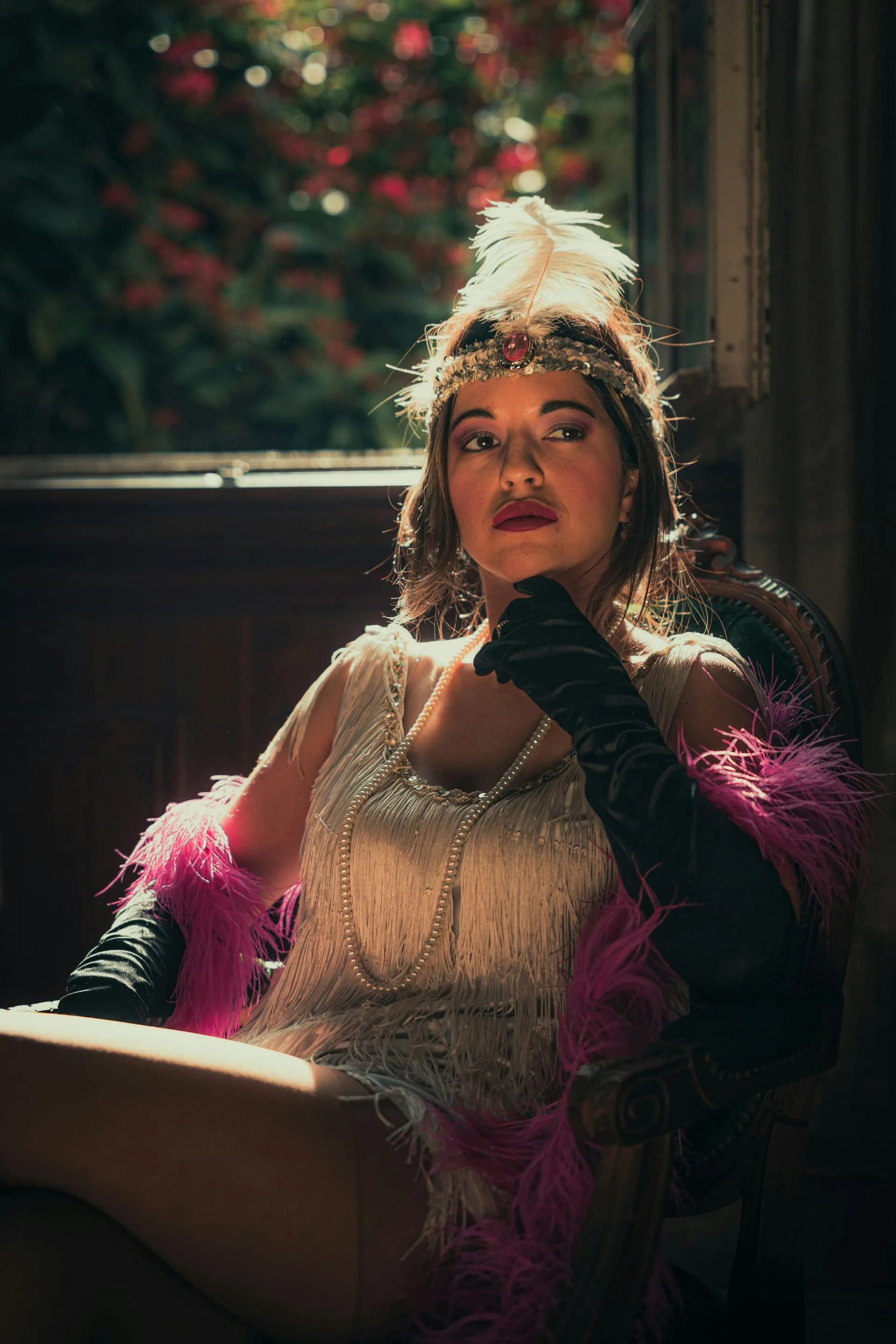 Woman in flapper dress, wearing a feathered headpiece and gloves, seated by a window, pink boa.
