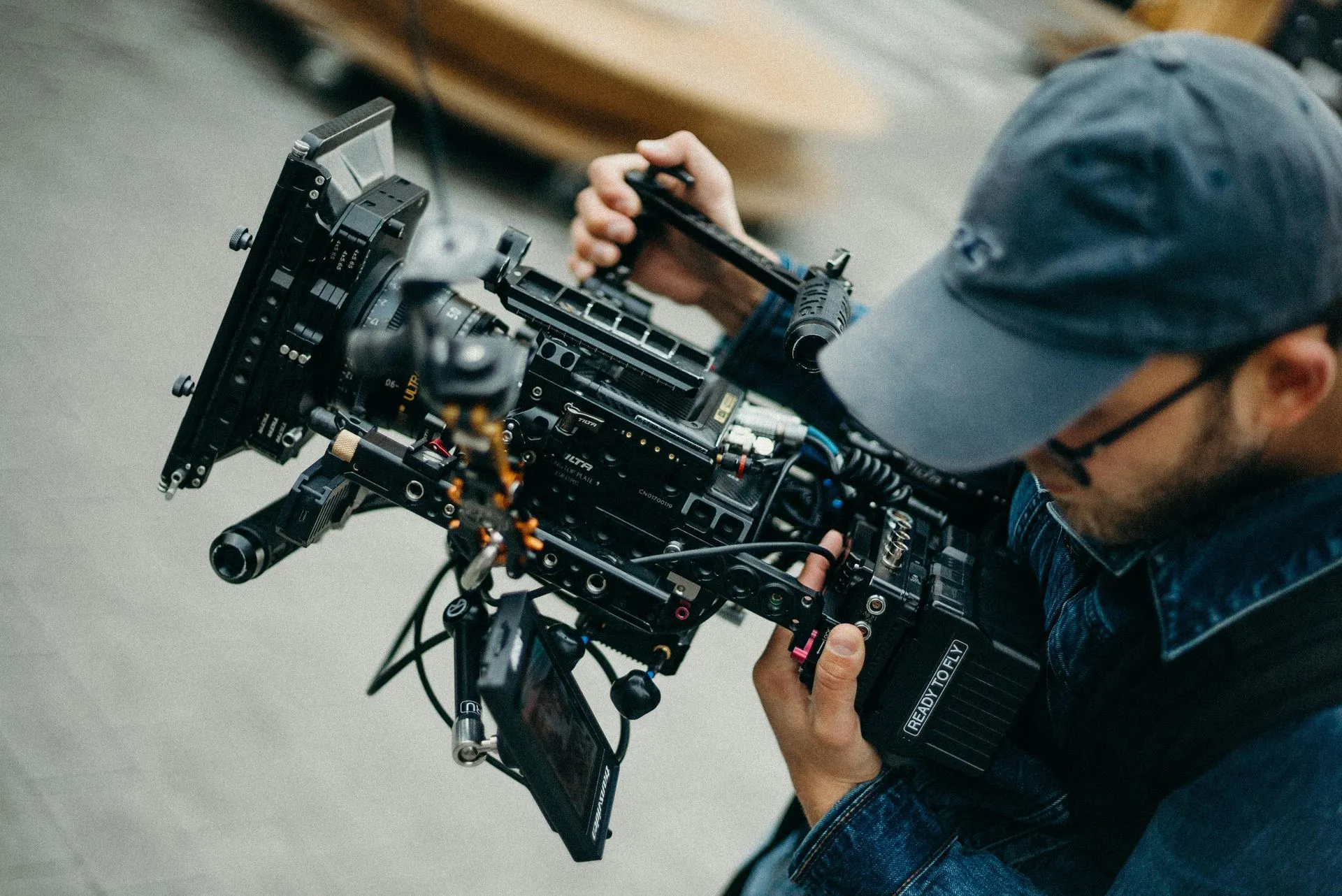 Person in denim jacket and cap holding a complex camera rig, focused, indoors.