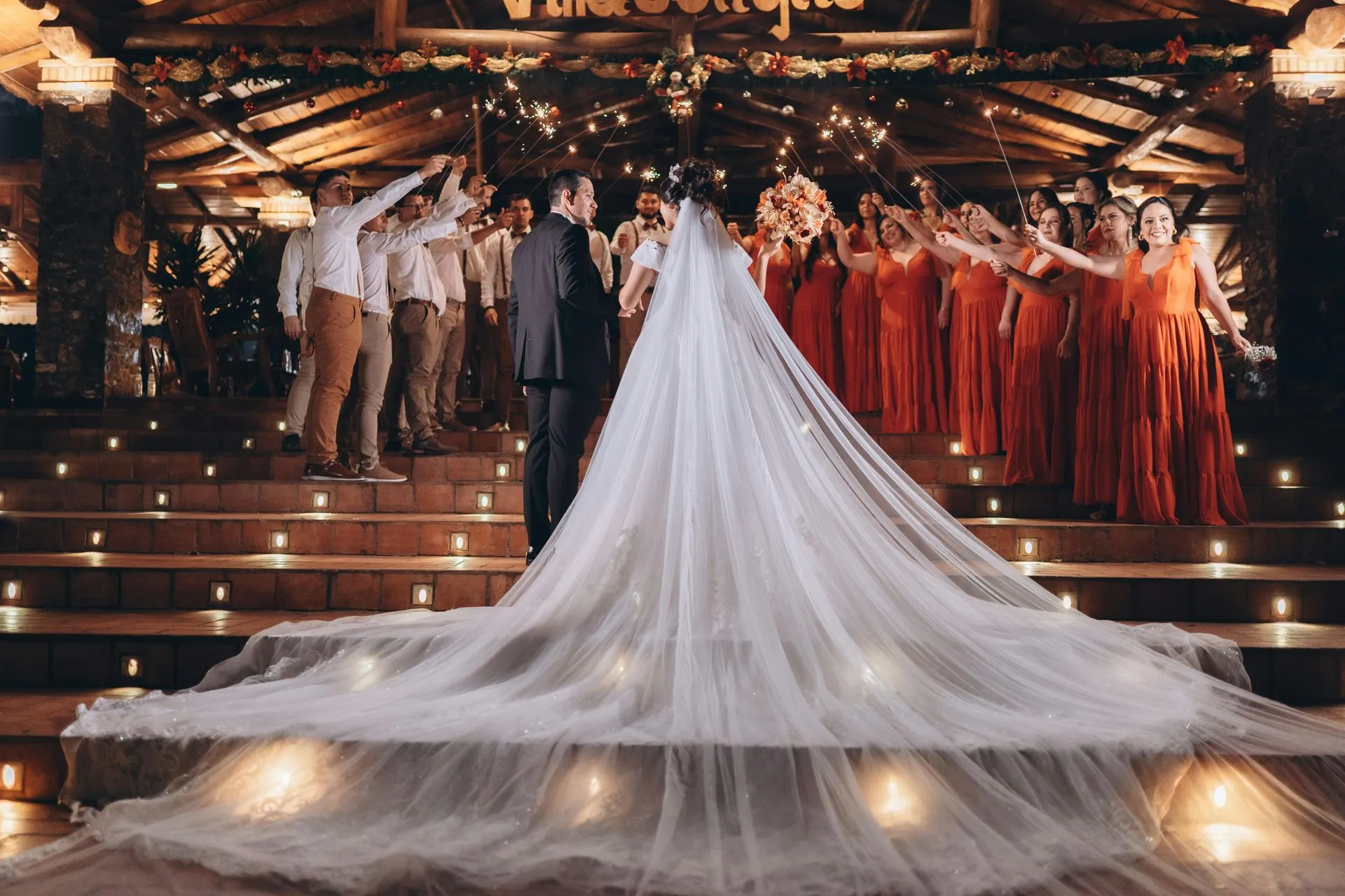 Wedding ceremony: Bride and groom on steps, veil trailing. Bridesmaids in red, groomsmen in white, lit setting.