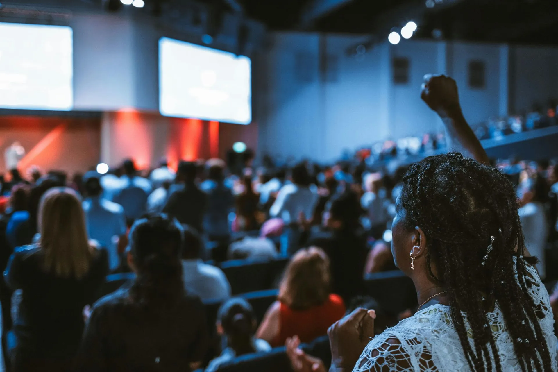 Person raising fist in crowded auditorium, possibly at a church service. Audience is blurred; screens visible.