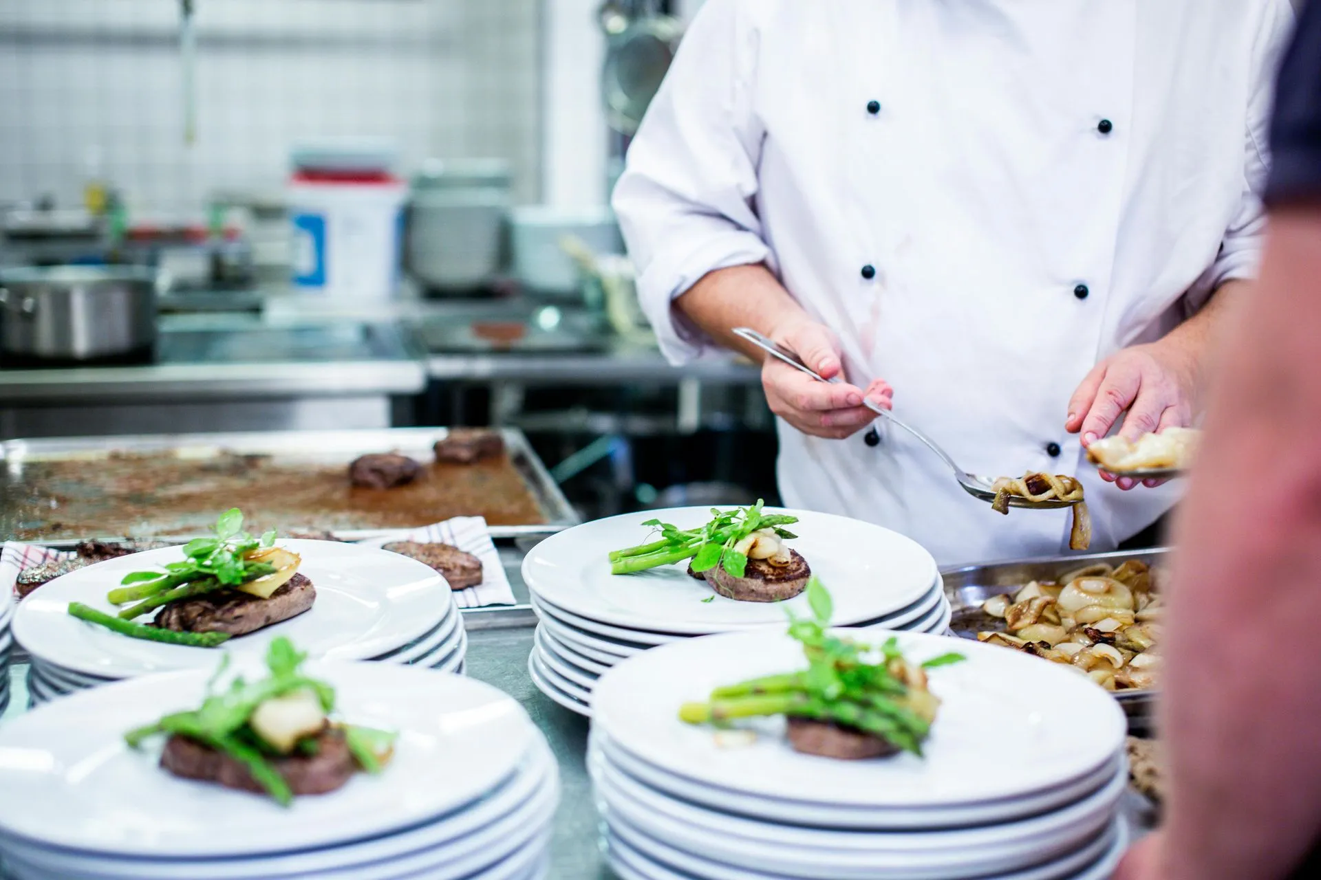 Chef plating dishes with steak, asparagus, and mushrooms in a commercial kitchen.