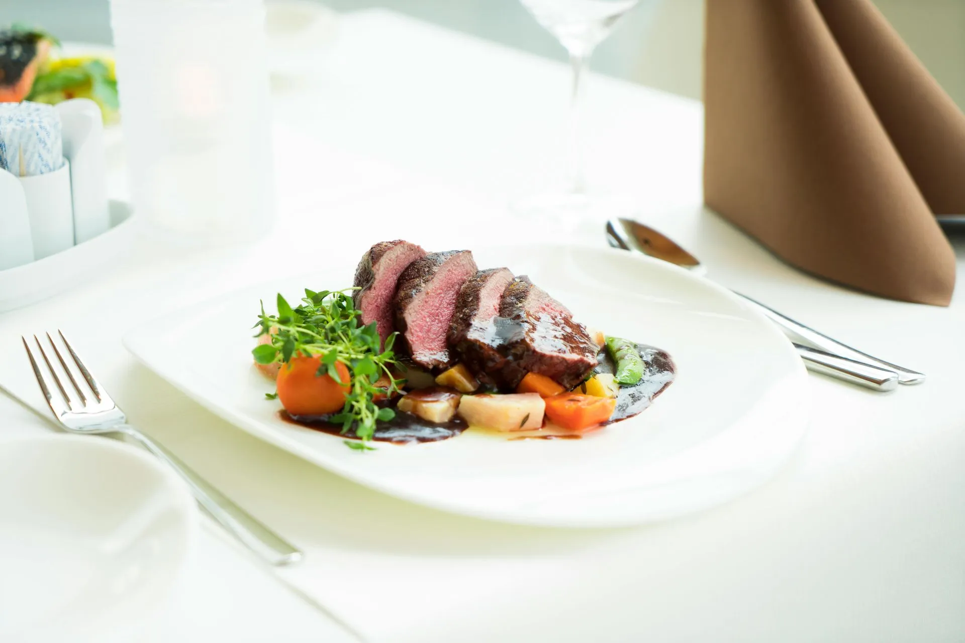Sliced steak with vegetables, herbs, and sauce on a white plate, set on a white tablecloth.