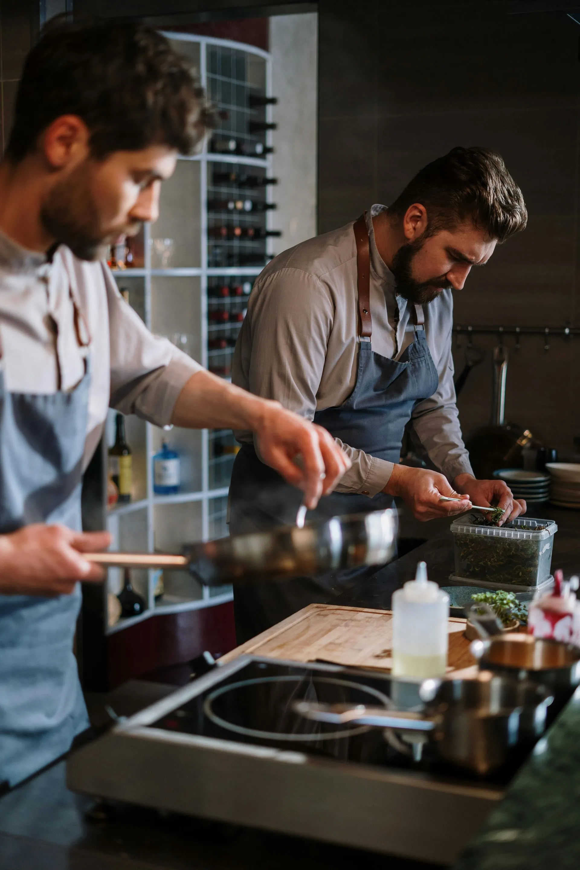 Two chefs in gray aprons cooking in a kitchen, one stirring a pan, the other preparing ingredients.
