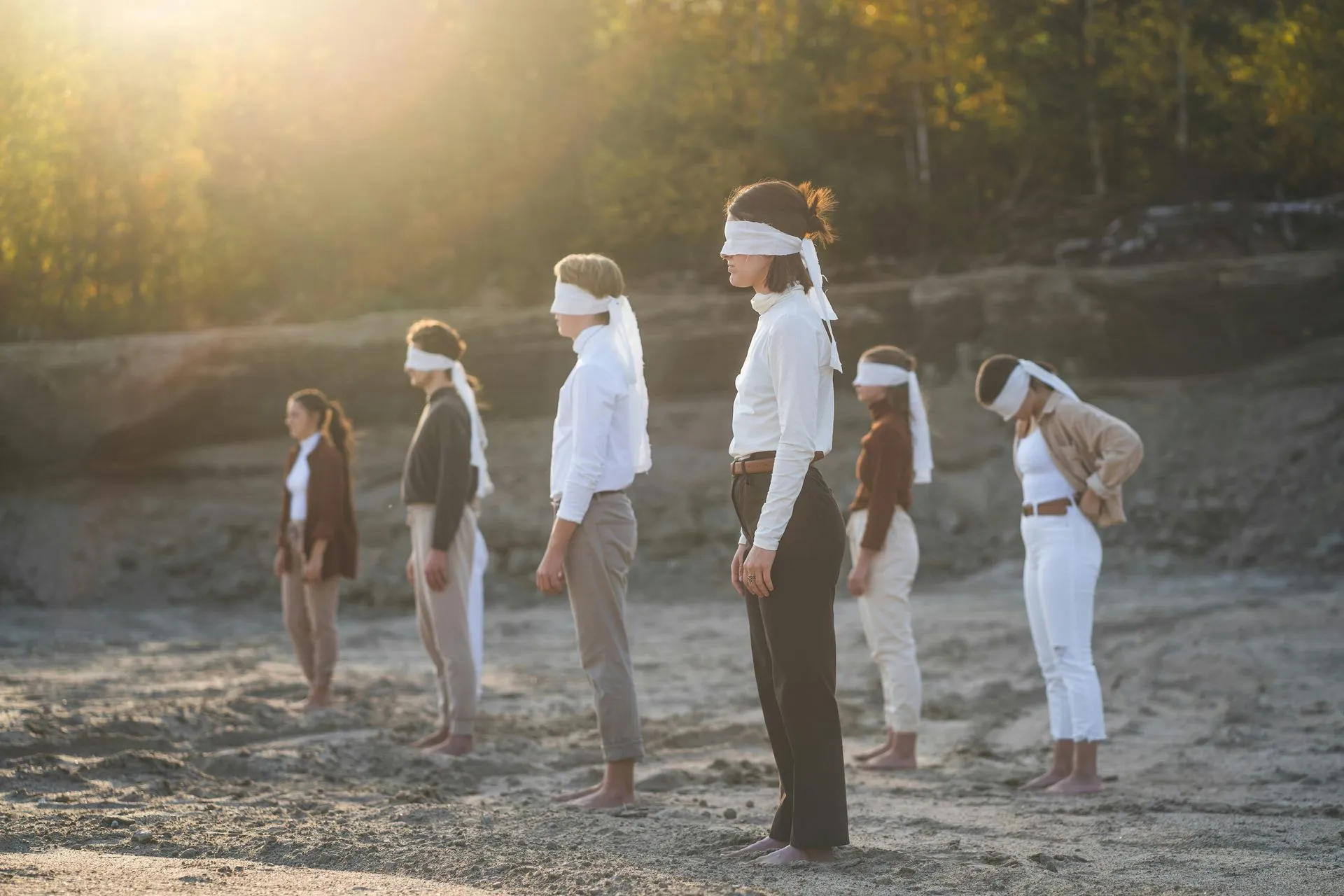 Seven people with white blindfolds stand on a gravelly surface, backlit by sunlight.