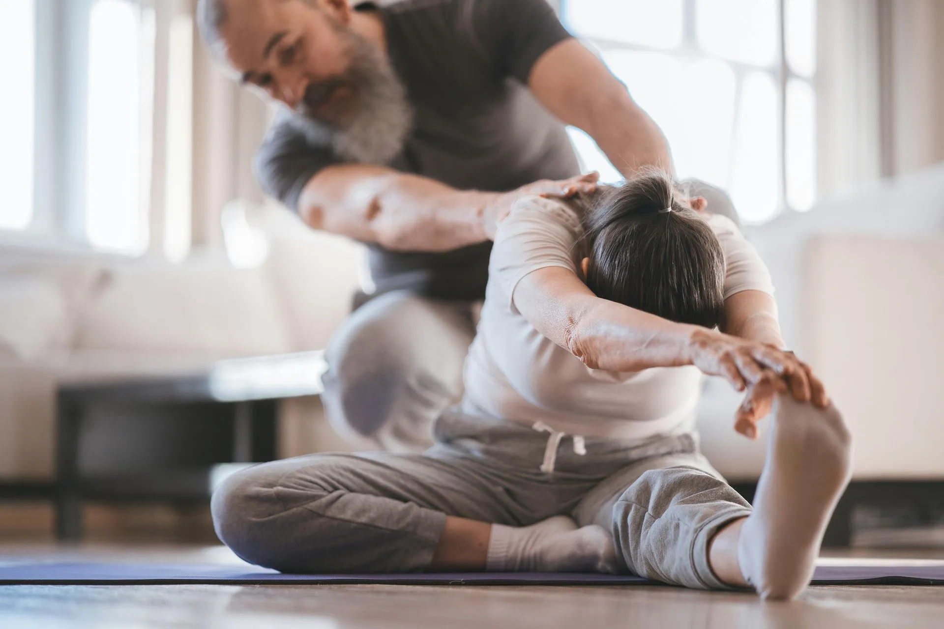 Man assisting person stretching leg on mat indoors.