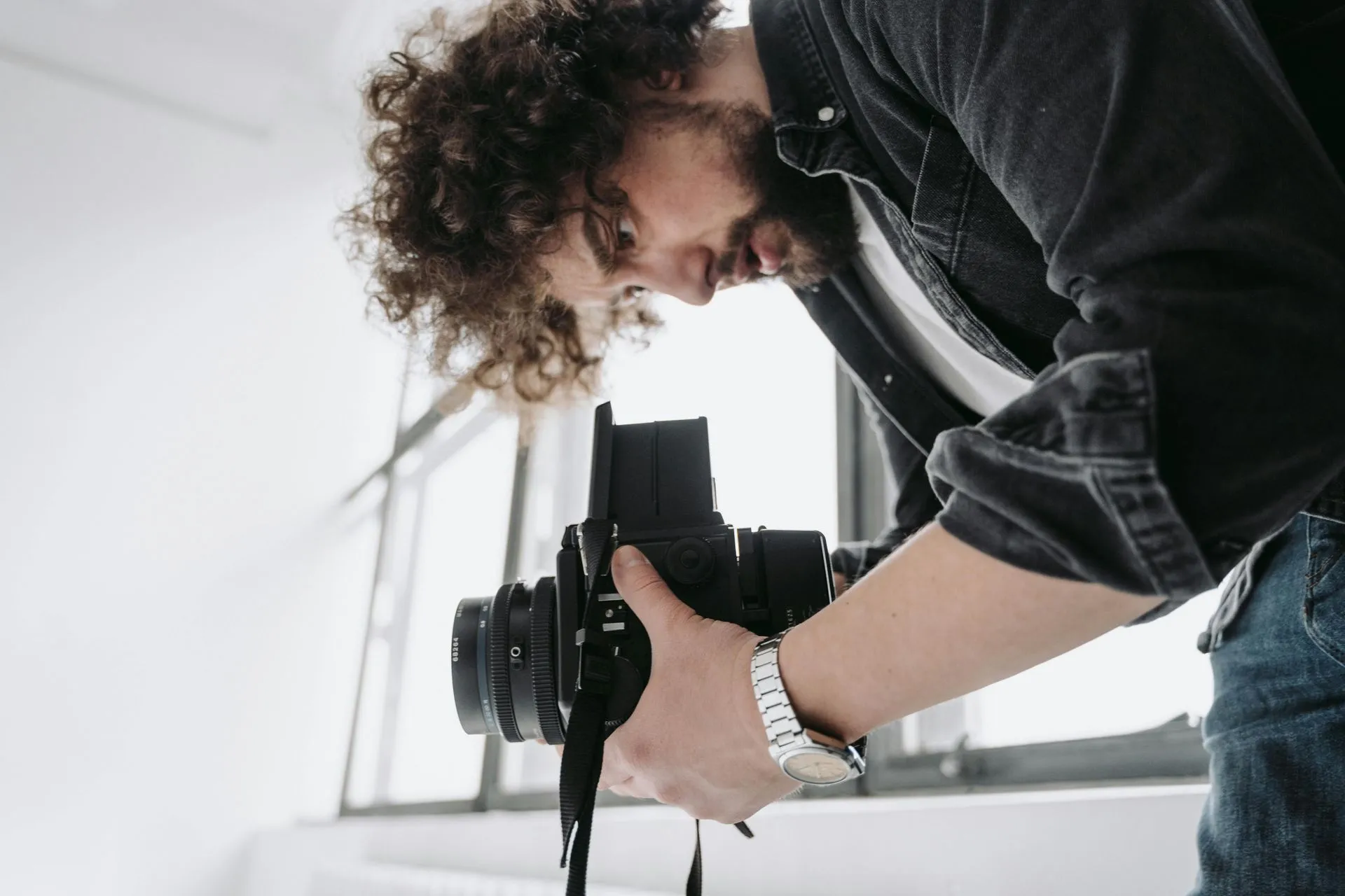 Man with curly hair, in denim shirt, looking through a camera lens, indoors with a white background.