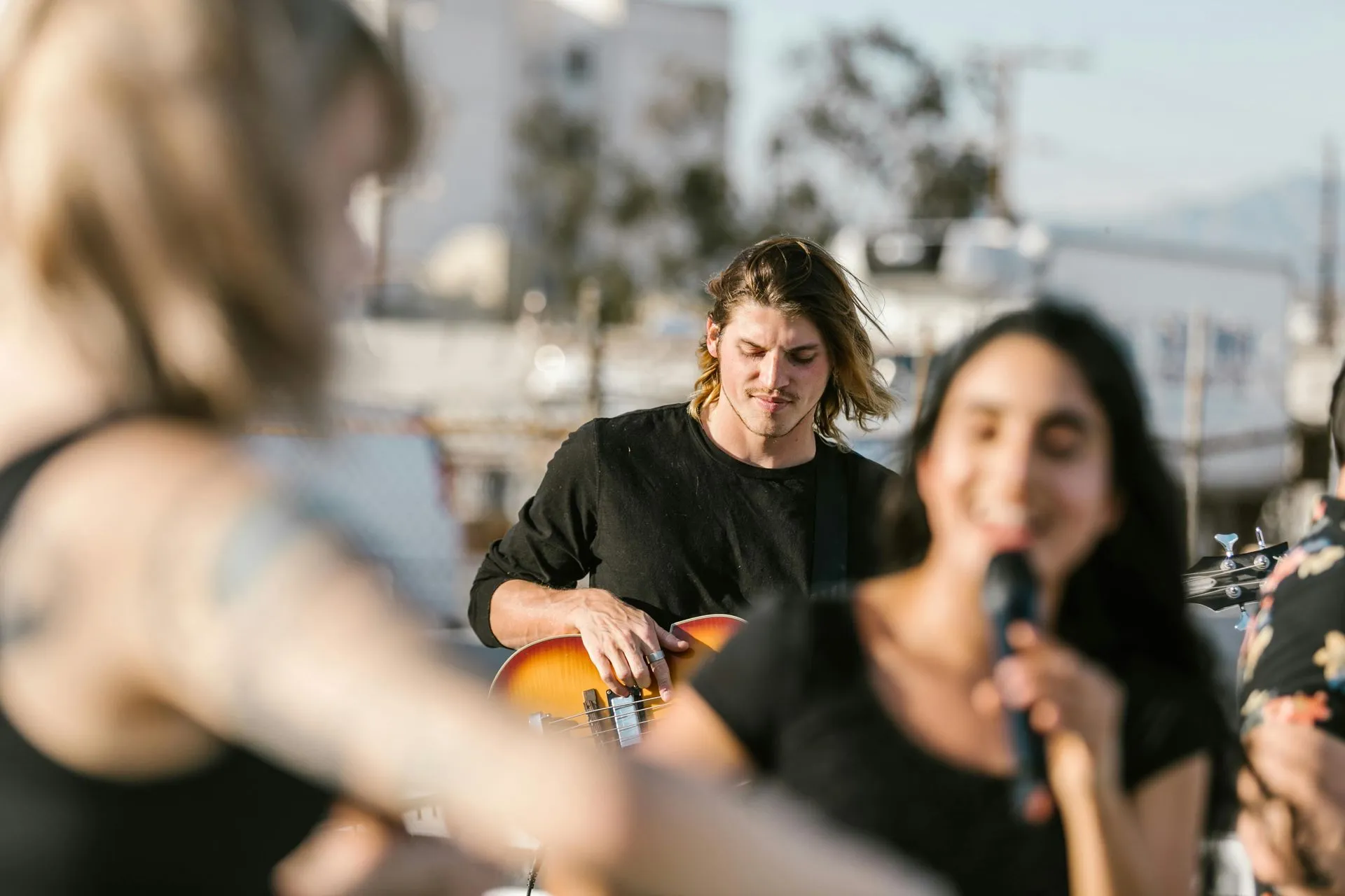 Band performing outdoors; guitarist in black shirt, singer with microphone smiling. Bright, sunny setting.