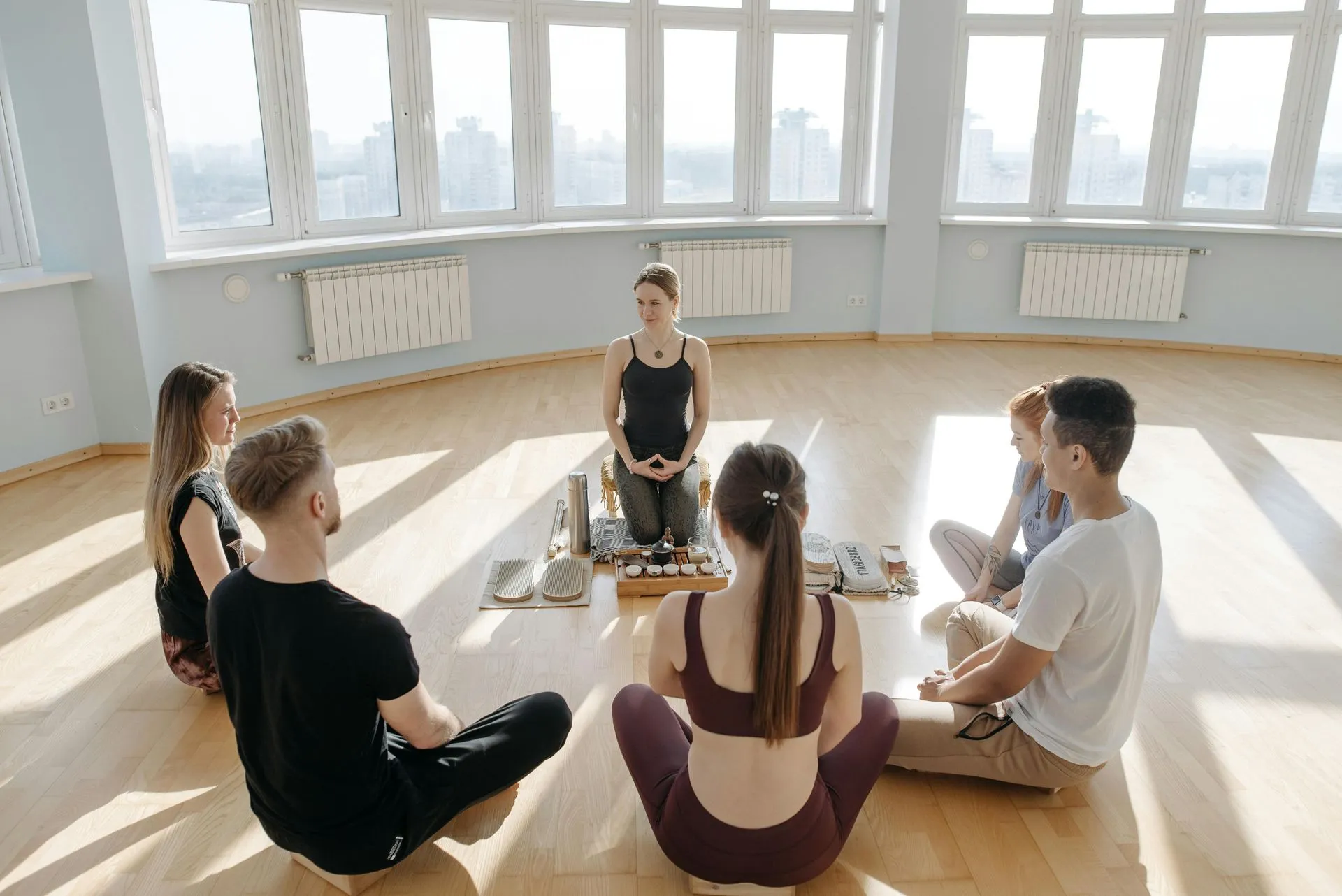 Group of people in a circle meditating around a woman in a light-filled room with wooden floors.