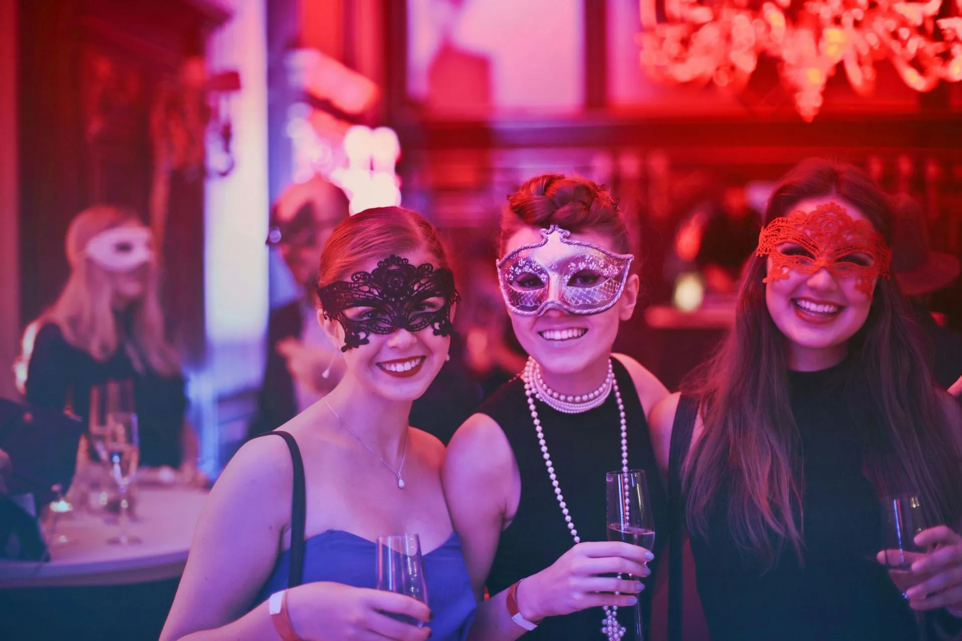 Three people wearing masks at a party, holding drinks and smiling. Lit with red and blue.