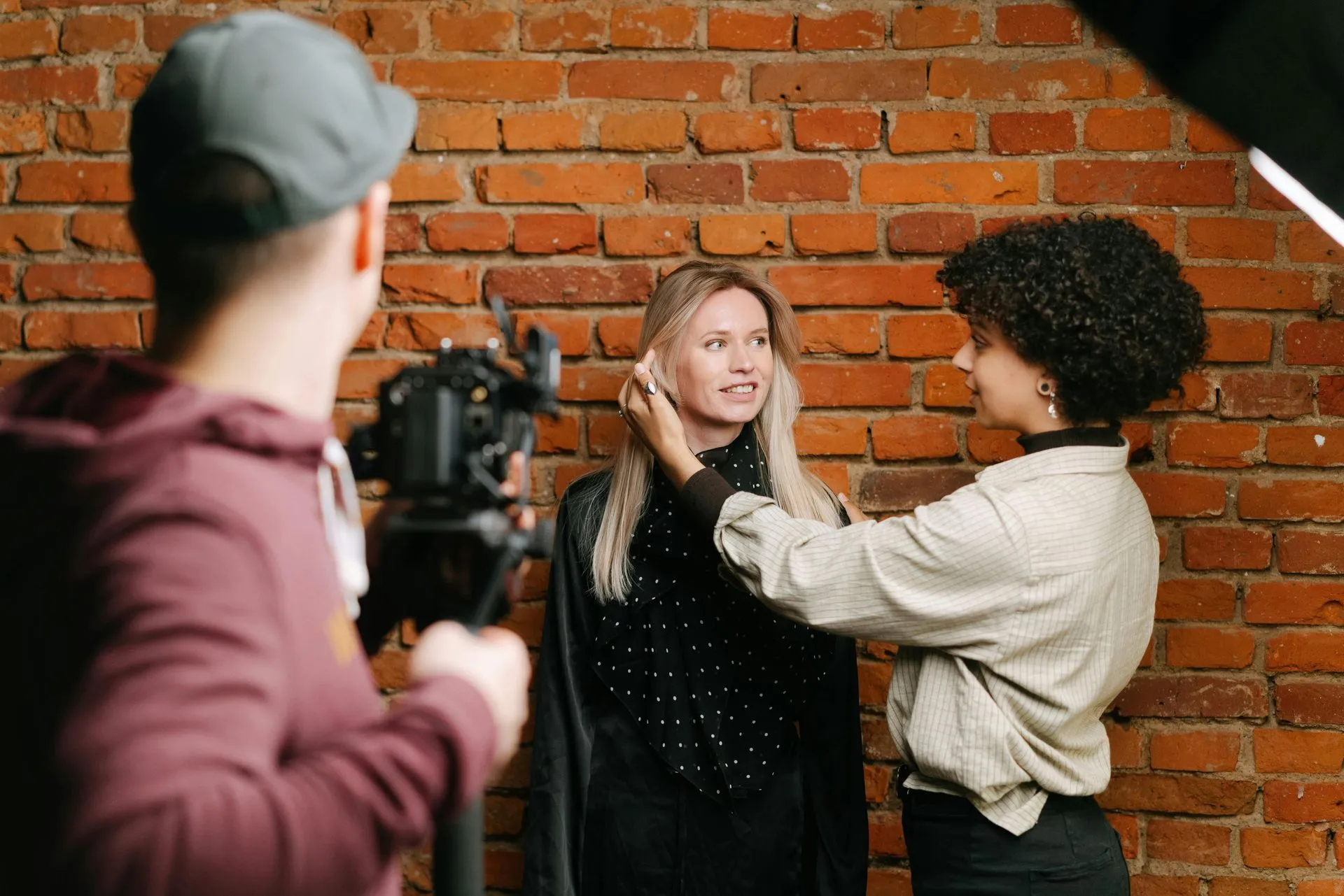 A photographer, makeup artist, and model pose against a brick wall. The makeup artist adjusts model's hair.