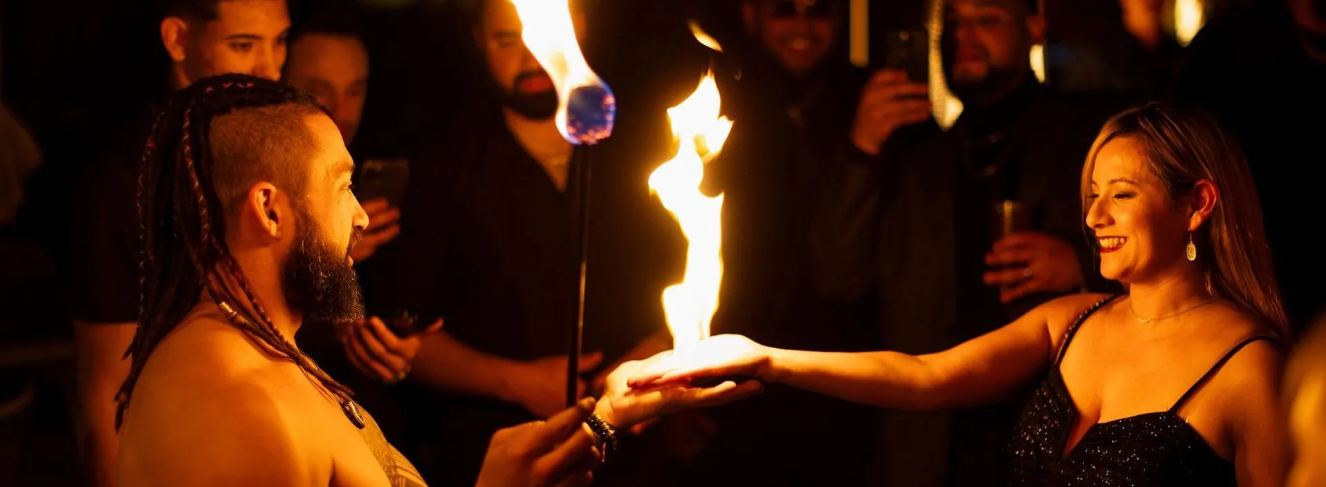 Two performers with fire poi at night, flames glowing, other spectators visible in darkened background watching