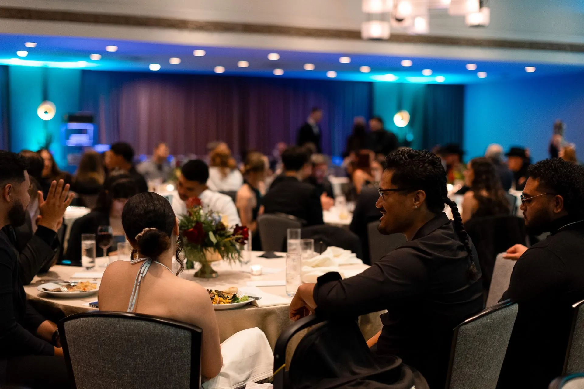 Guests in formal wear seated at round tables with place settings under blue lighting