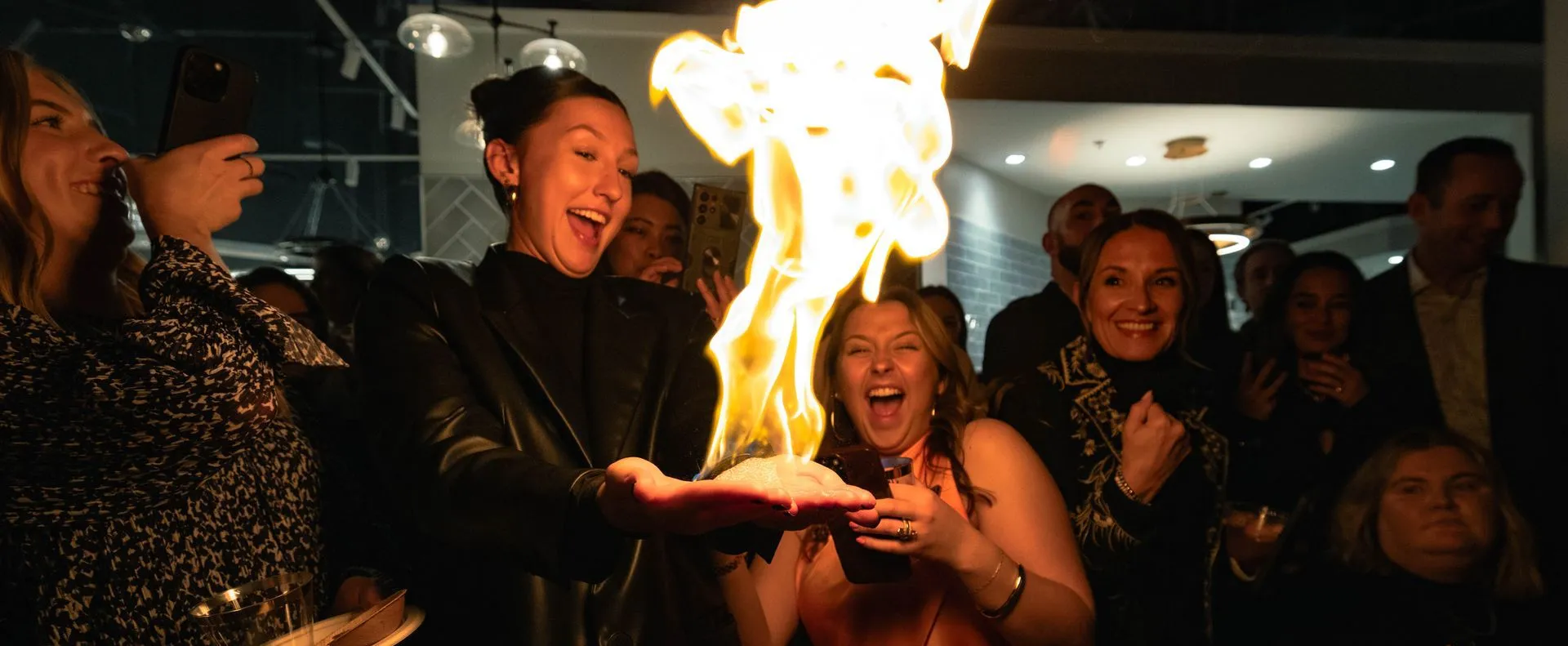Fire performer spinning flames from hands surrounded by spectators in formal attire at indoor event